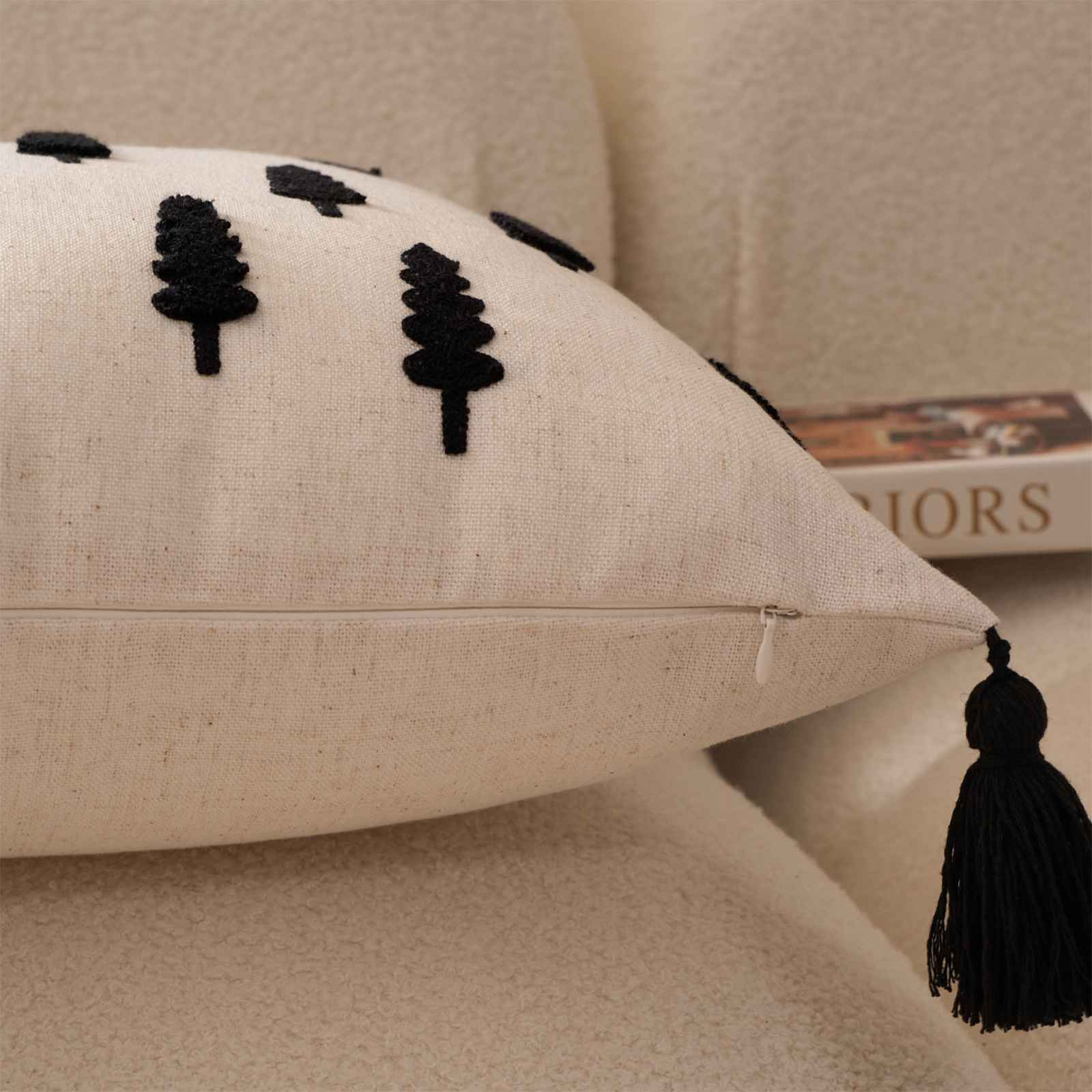 Cremefarbenes Kissen mit schwarzen Baumdesigns, Reißverschluss und Quaste. Es liegt auf einem beigen Sofa.