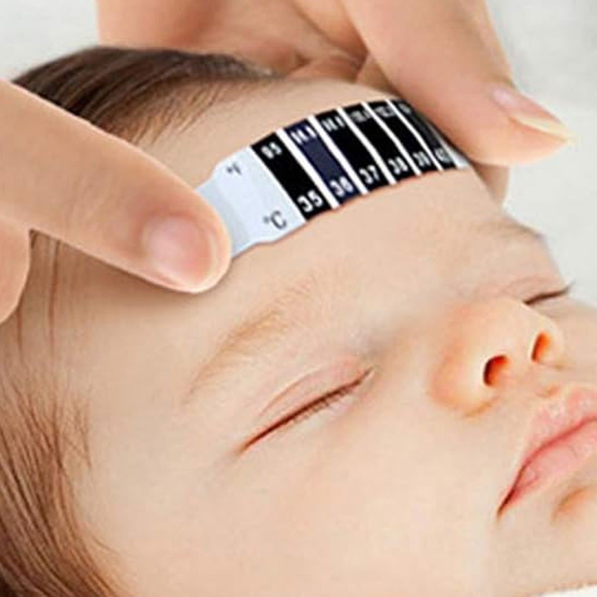 Eine Hand hält ein Stirnthermometer auf der Stirn eines Babys. Das Thermometer zeigt Temperaturwerte an. Die Augen des Babys sind geschlossen.