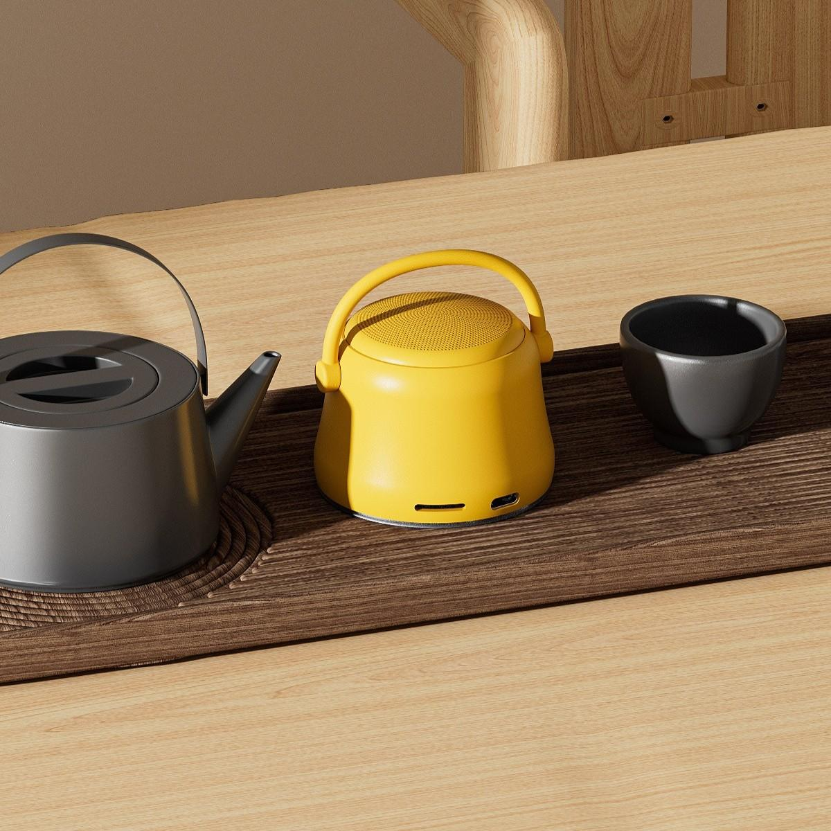Ein gelber Lautsprecher mit Griff, eine graue Teekanne und eine schwarze Teetasse auf einem Holztisch.