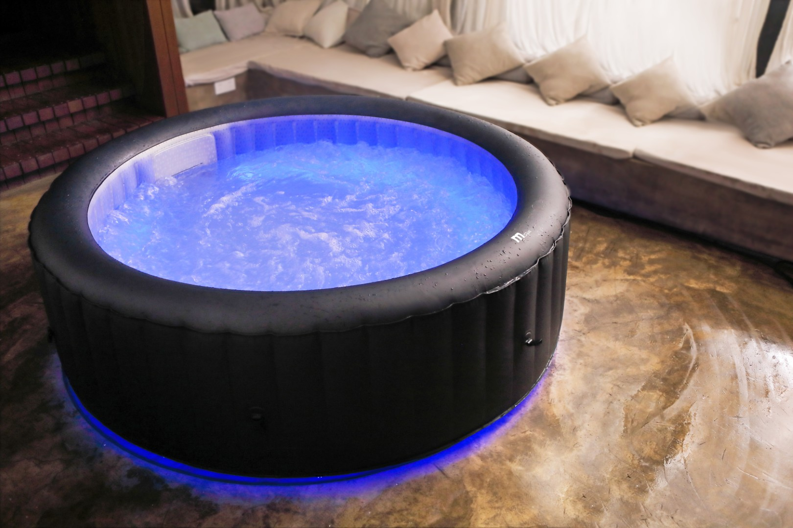 Ein runder, schwarzer, aufblasbarer Whirlpool mit blauem Wasser und Lichtern. Kissen und eine Bank sind im Hintergrund.