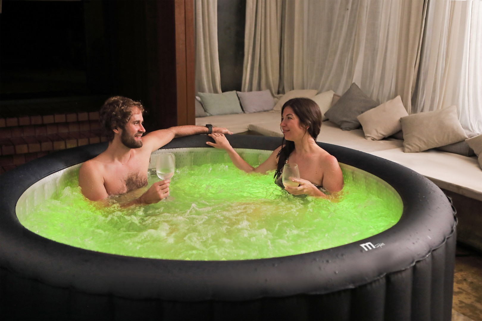 Ein Paar genießt einen Whirlpool mit grünem Wasser und hält Weingläser. Im Hintergrund Vorhänge und eine Sitzecke mit Kissen.