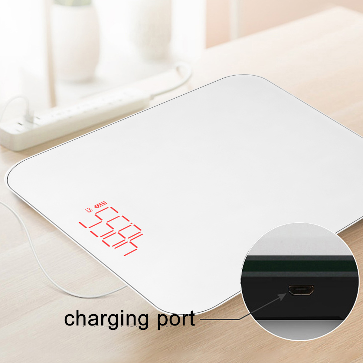 Weiße Digitalwaage mit 55,4 kg. Ein Ladeanschluss befindet sich an der Seite. Eine Steckdosenleiste und Vase sind im unscharfen Hintergrund.