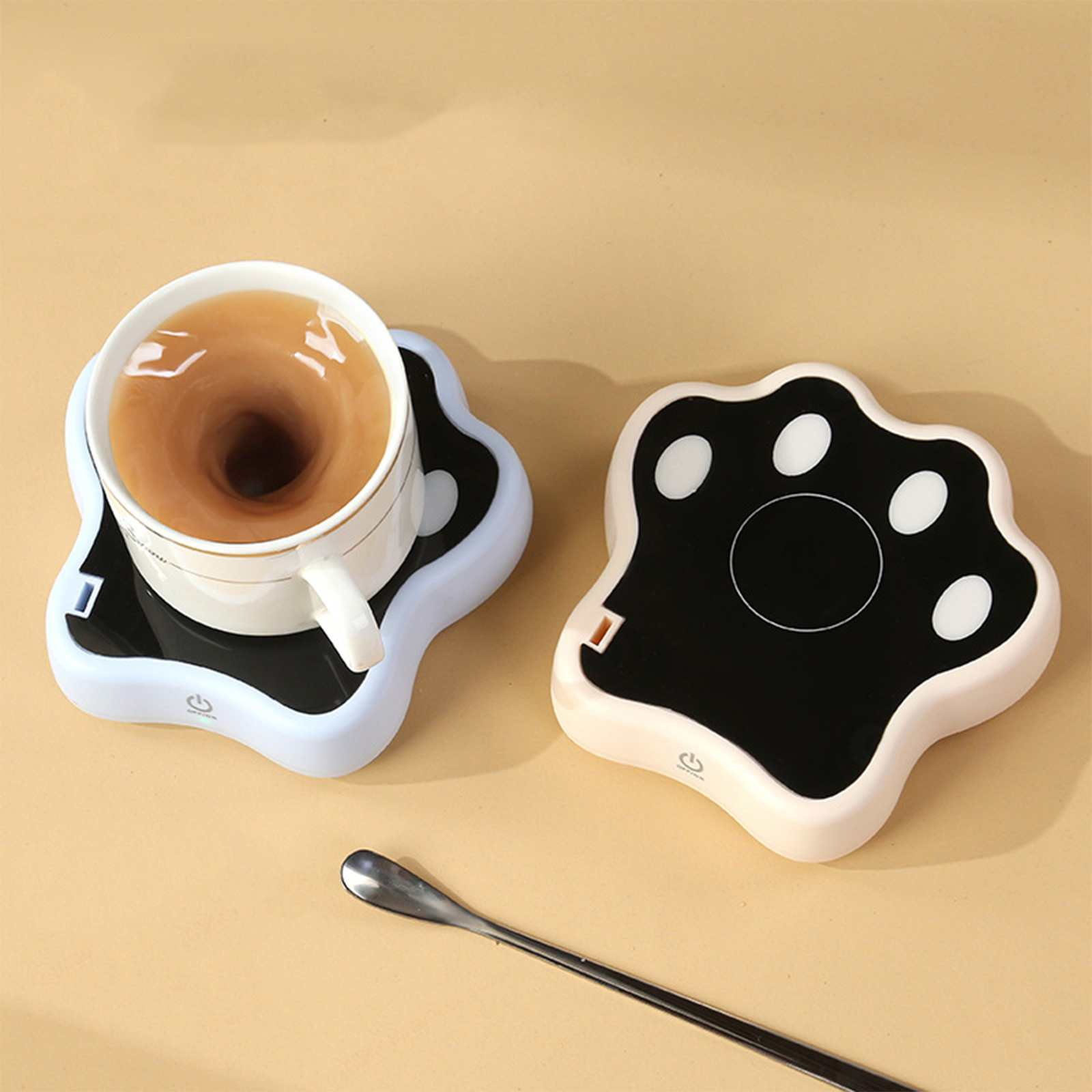 Zwei pfotenförmige Tassenwärmer, einer mit einer Kaffeetasse. Ein Löffel ist in der Nähe. Beigefarbener Hintergrund.