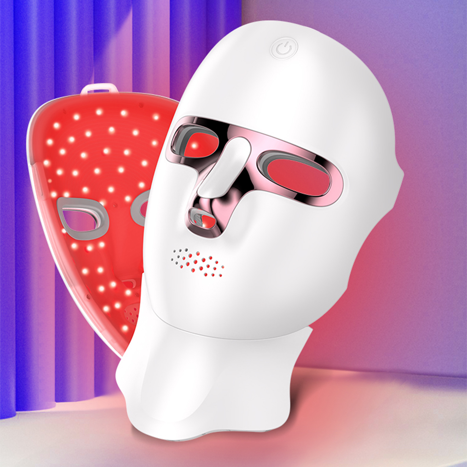 L'immagine mostra una maschera facciale a LED bianca con una maschera a LED rossa dietro, su uno sfondo di tende viola e blu.