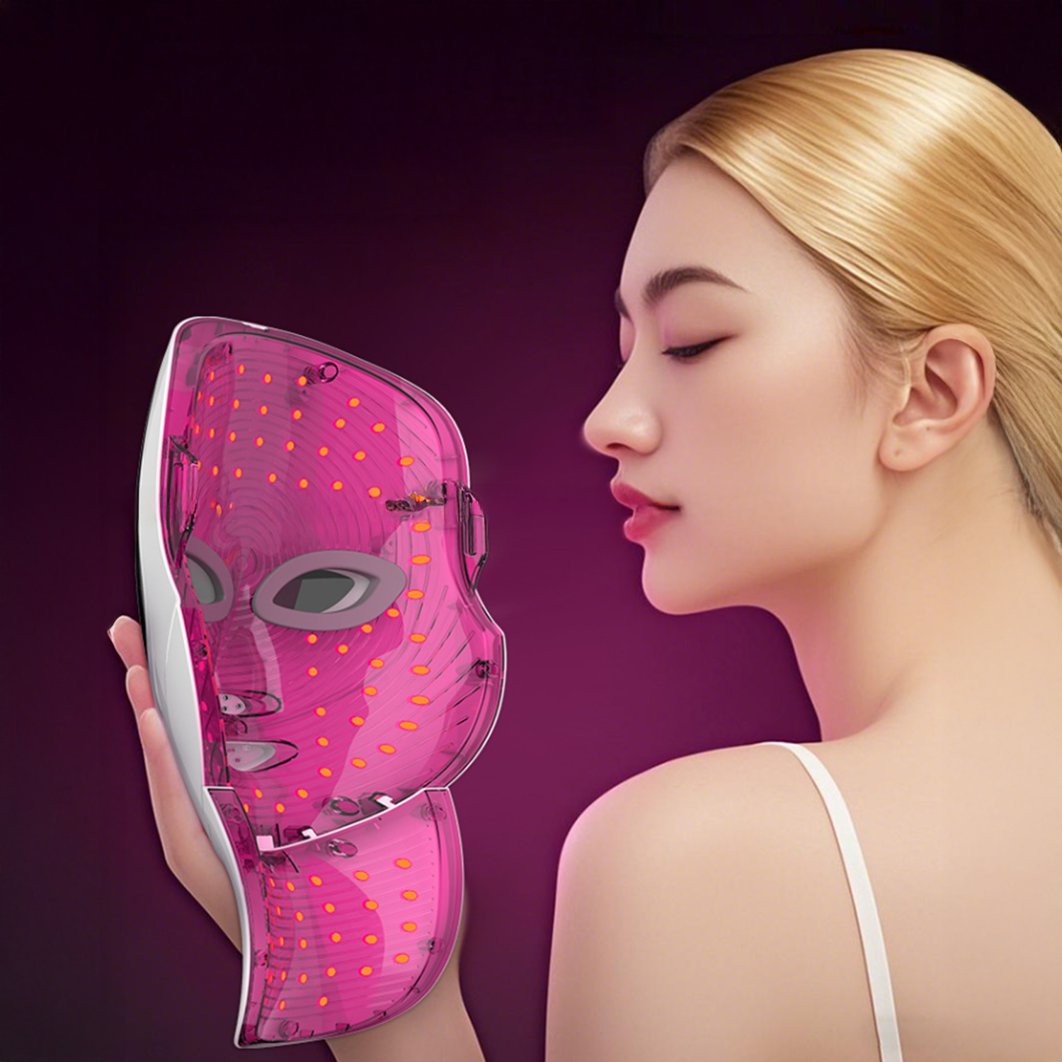 Eine Frau hält eine rosa LED-Gesichtsmaske. Sie hat blondes Haar und geschlossene Augen. Der Hintergrund ist dunkelviolett.