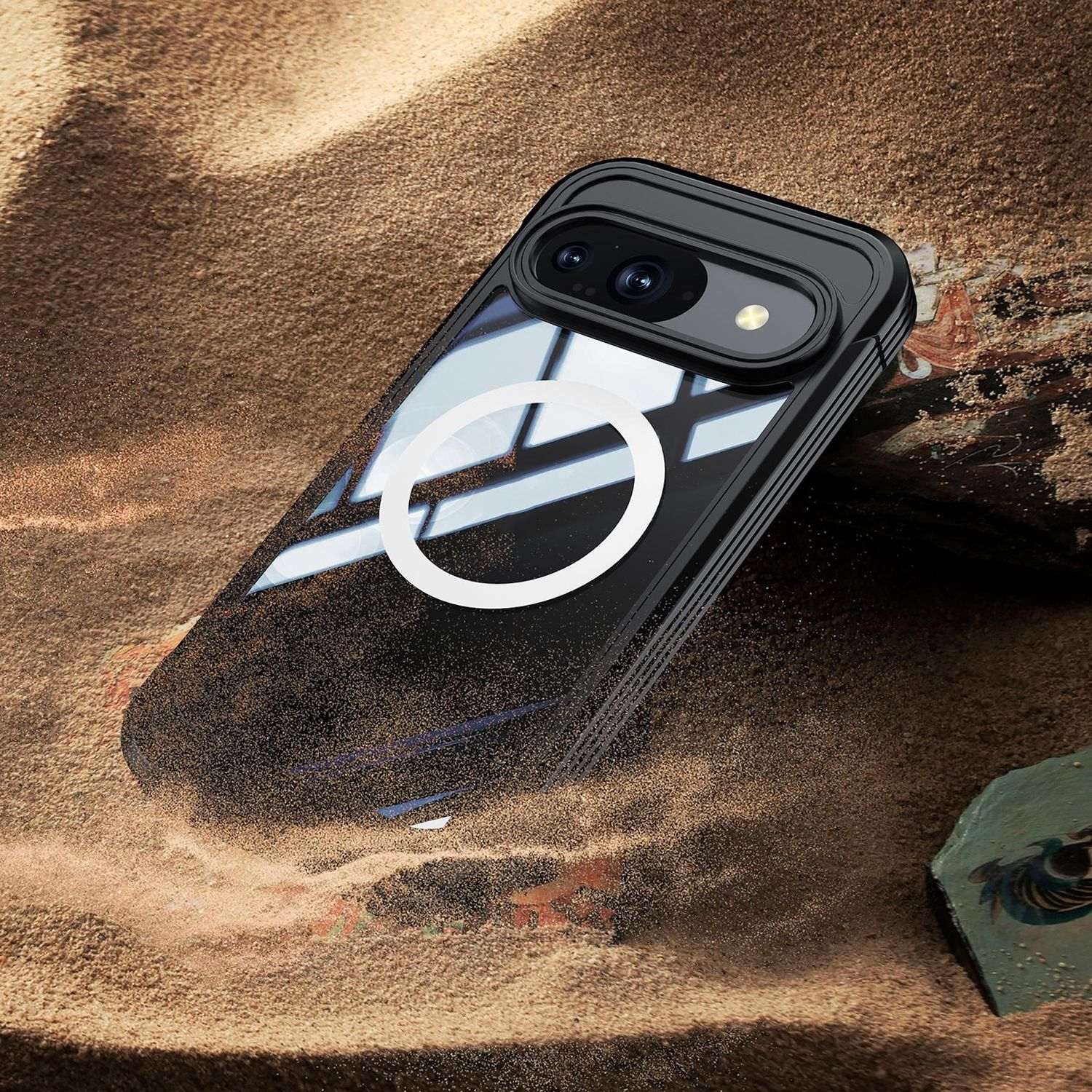 Eine schwarze Handyhülle mit weißem Ring liegt im Sand. Die Hülle hat eine Kameraerhöhung. Sand umgibt die Hülle, und ein Stein liegt im Sand.