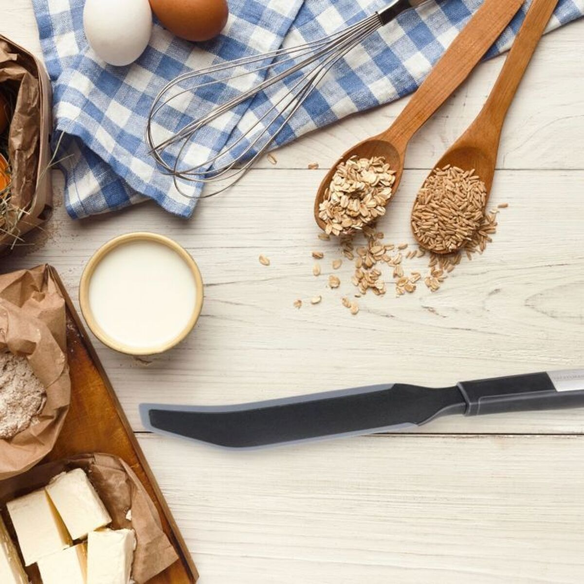 Draufsicht auf Backzutaten: Eier, Milch, Hafer, Butter, Mehl und ein Messer auf einem Holztisch mit blauem Karotuch.