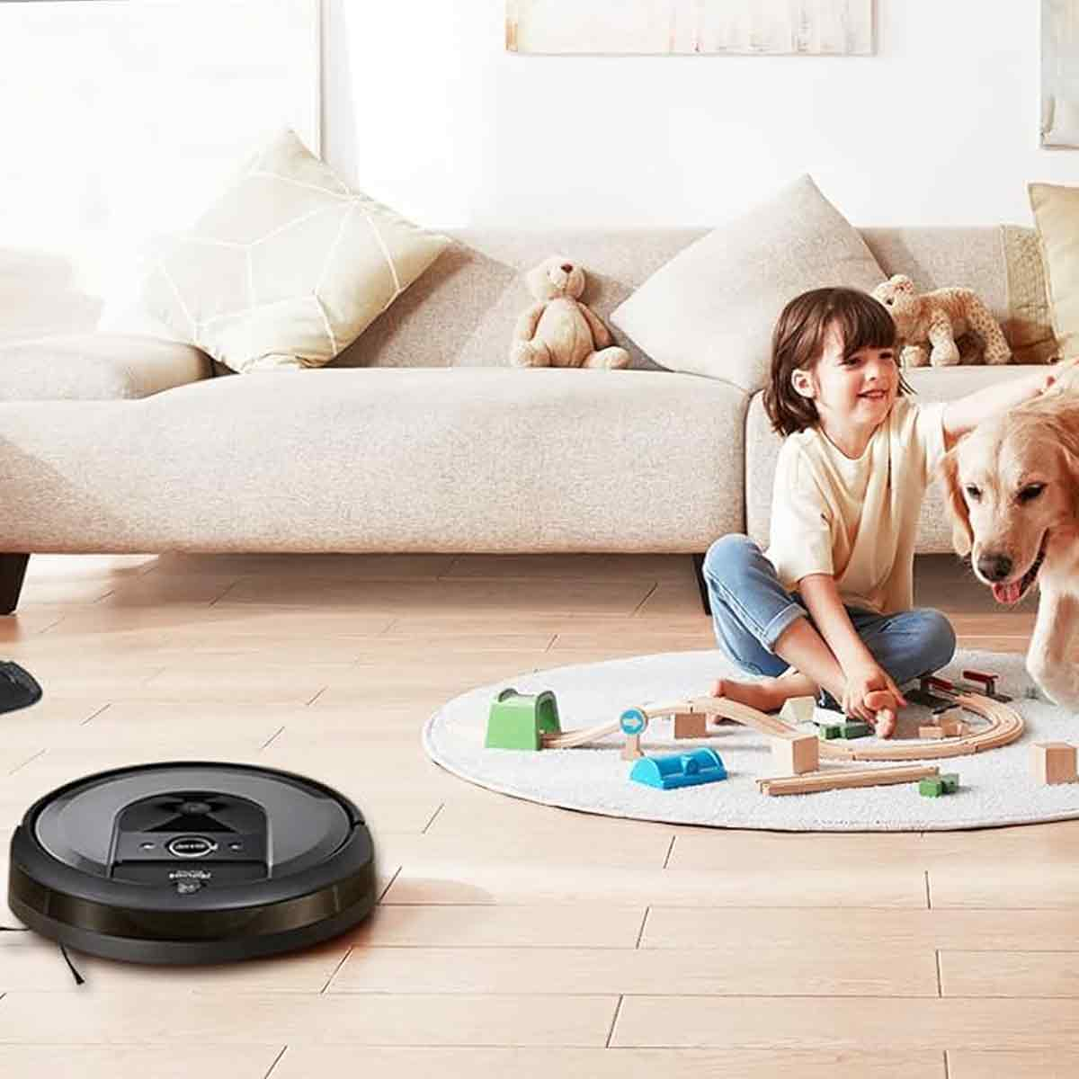 Ein Mädchen sitzt mit einem Hund in der Nähe eines Spielzeugeisenbahn-Sets auf einem Teppich. Ein Roboter-Staubsauger steht auf dem Holzboden. Im Hintergrund ist ein Sofa.