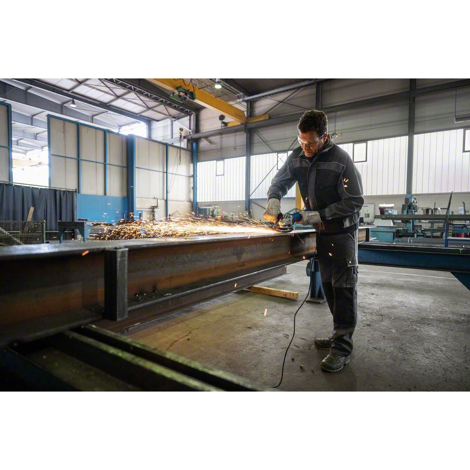 Ein Mann schleift einen Metallträger mit einem Schleifer, Funken fliegen. Er trägt Schutzkleidung, in einer Werkstatt.