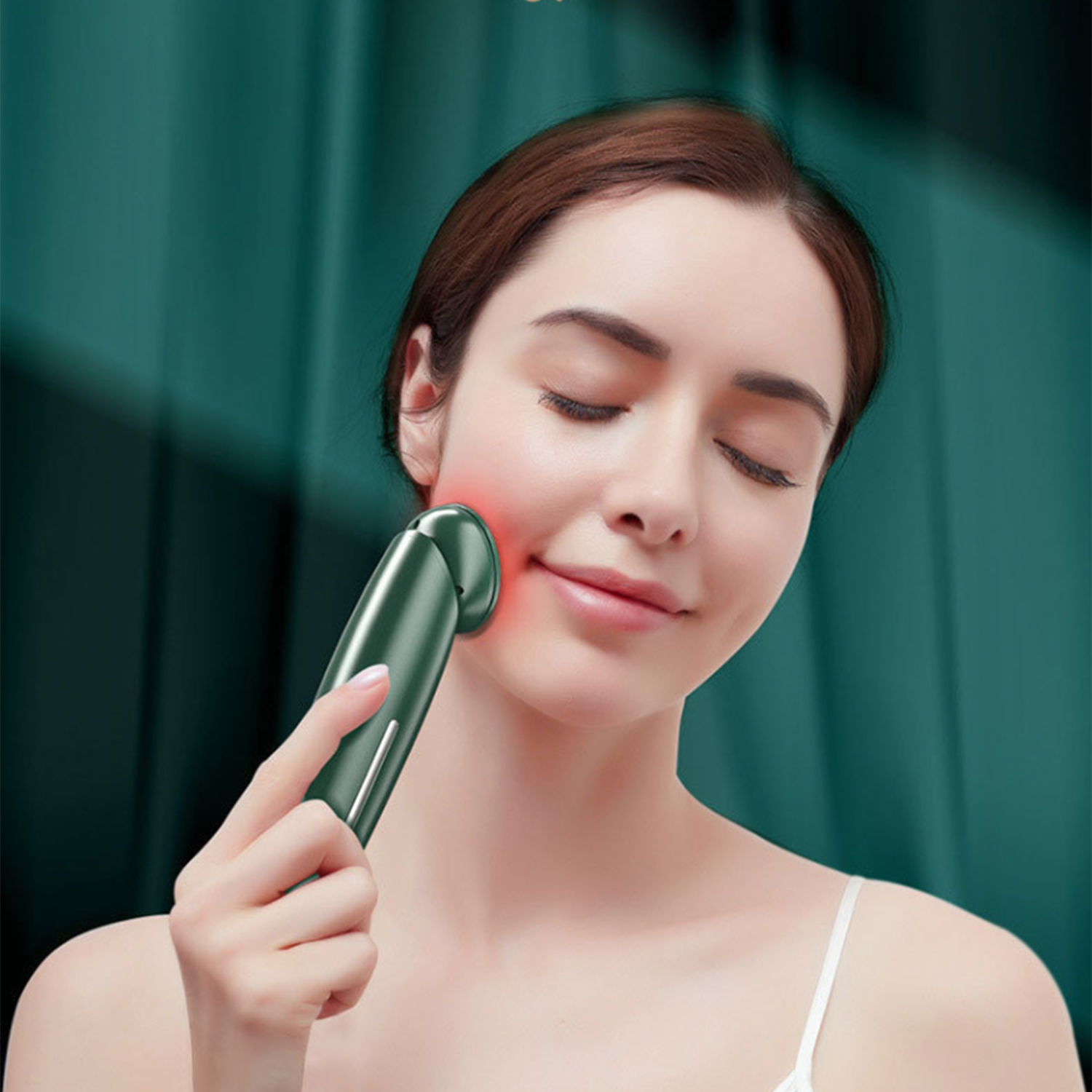 Una mujer con los ojos cerrados usa un masajeador facial verde. Luz roja en su rostro. Viste un top blanco ante una cortina verde.