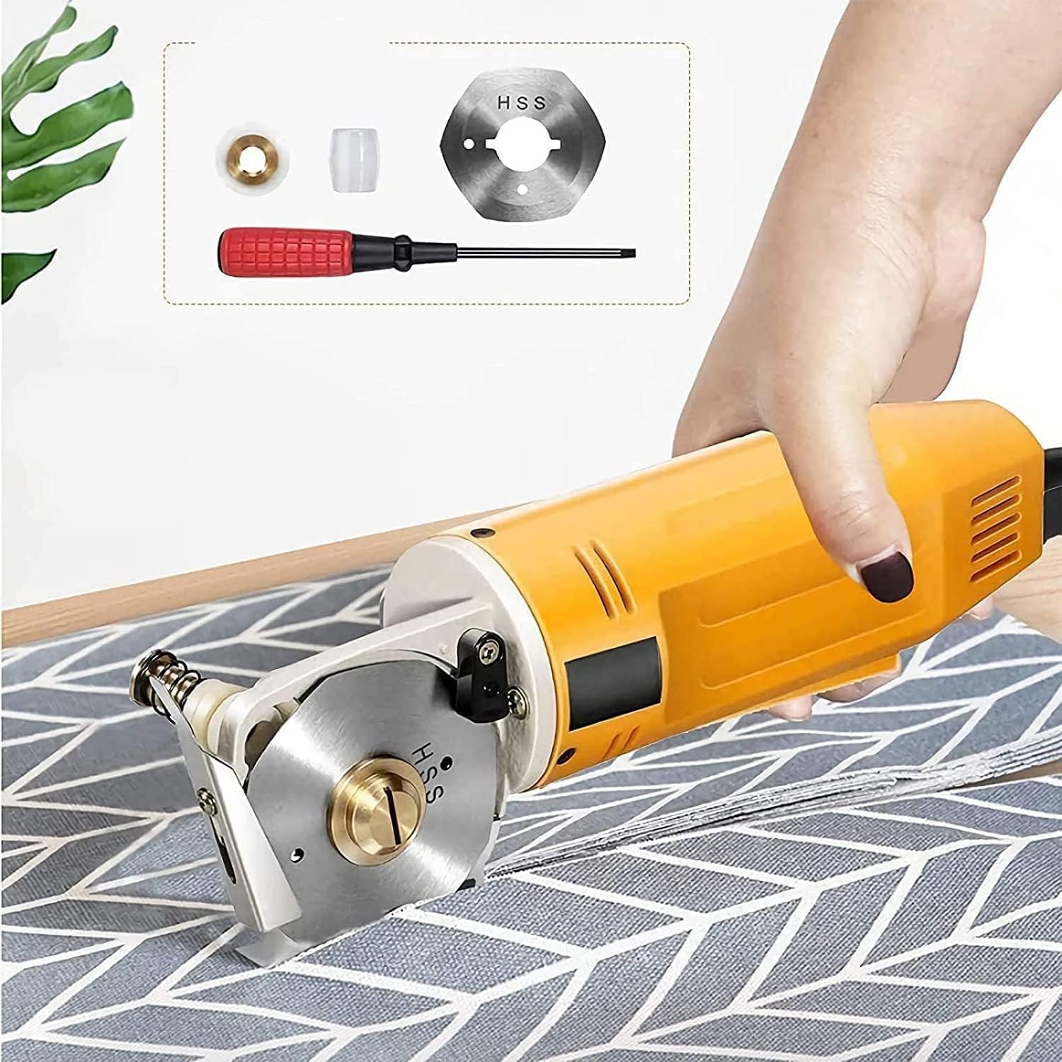Eine Hand hält einen orangefarbenen elektrischen Stoffschneider. Ersatzteile sind oben. Der Schneider schneidet einen grauen Stoff mit Muster auf einem Holztisch.
