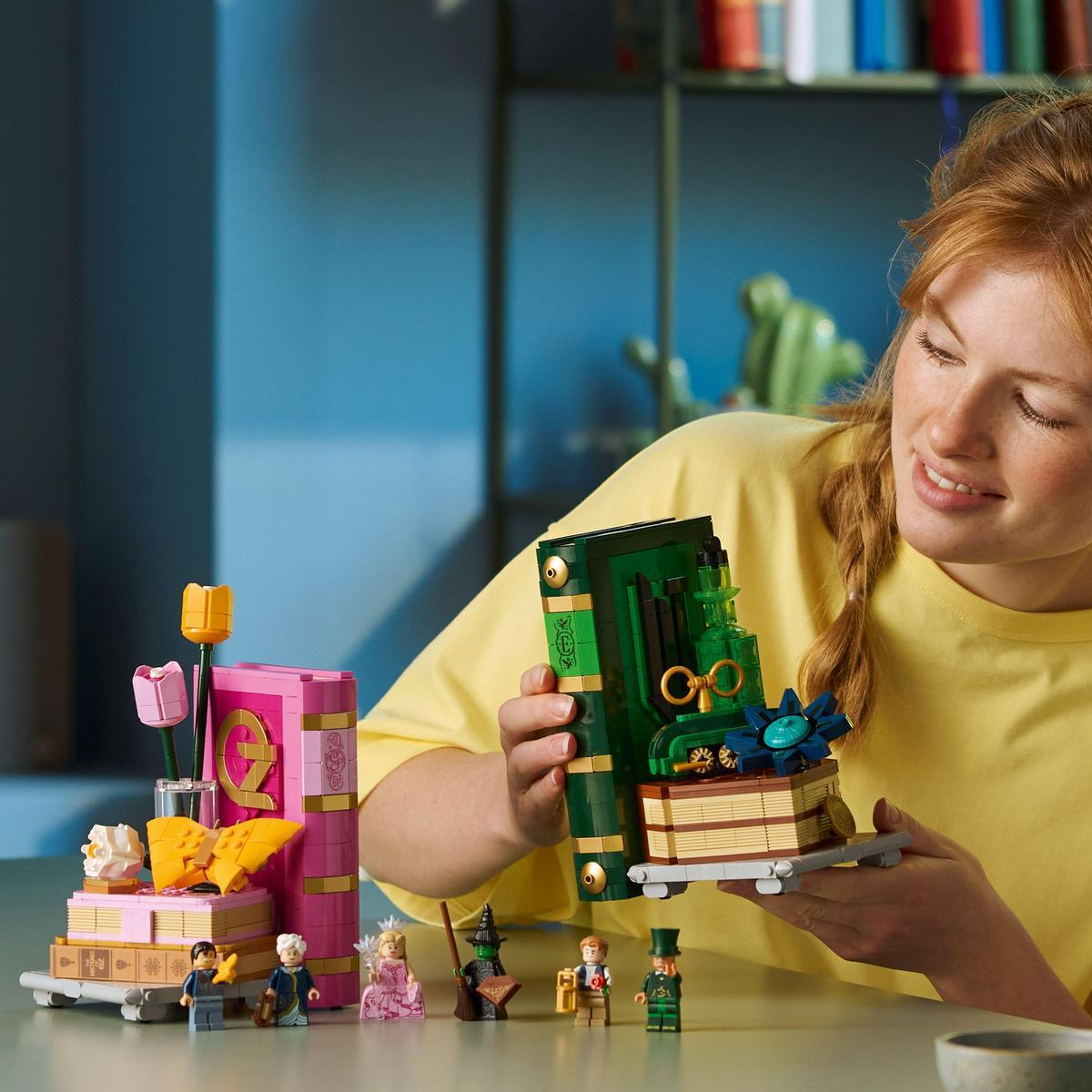 Een vrouw houdt een Lego-boek vast. Lego bloemen en figuren staan op een tafel. De scène bevat groene, roze, gele en blauwe elementen.