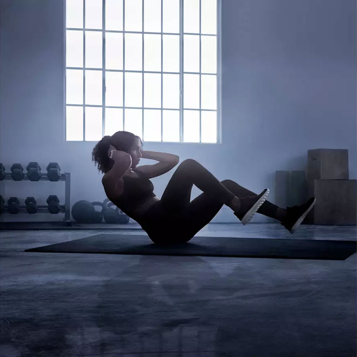 Frau in schwarzer Sportkleidung macht Sit-ups auf einer Matte. Im Hintergrund ist ein Fenster zu sehen, Gewichte sind sichtbar.