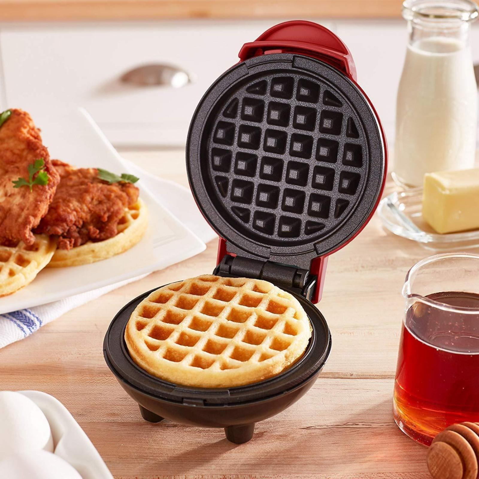 Ein Waffeleisen mit einer fertigen Waffel. Gebratenes Hähnchen und Sirup sind in der Nähe. Butter und Milch sind ebenfalls vorhanden.