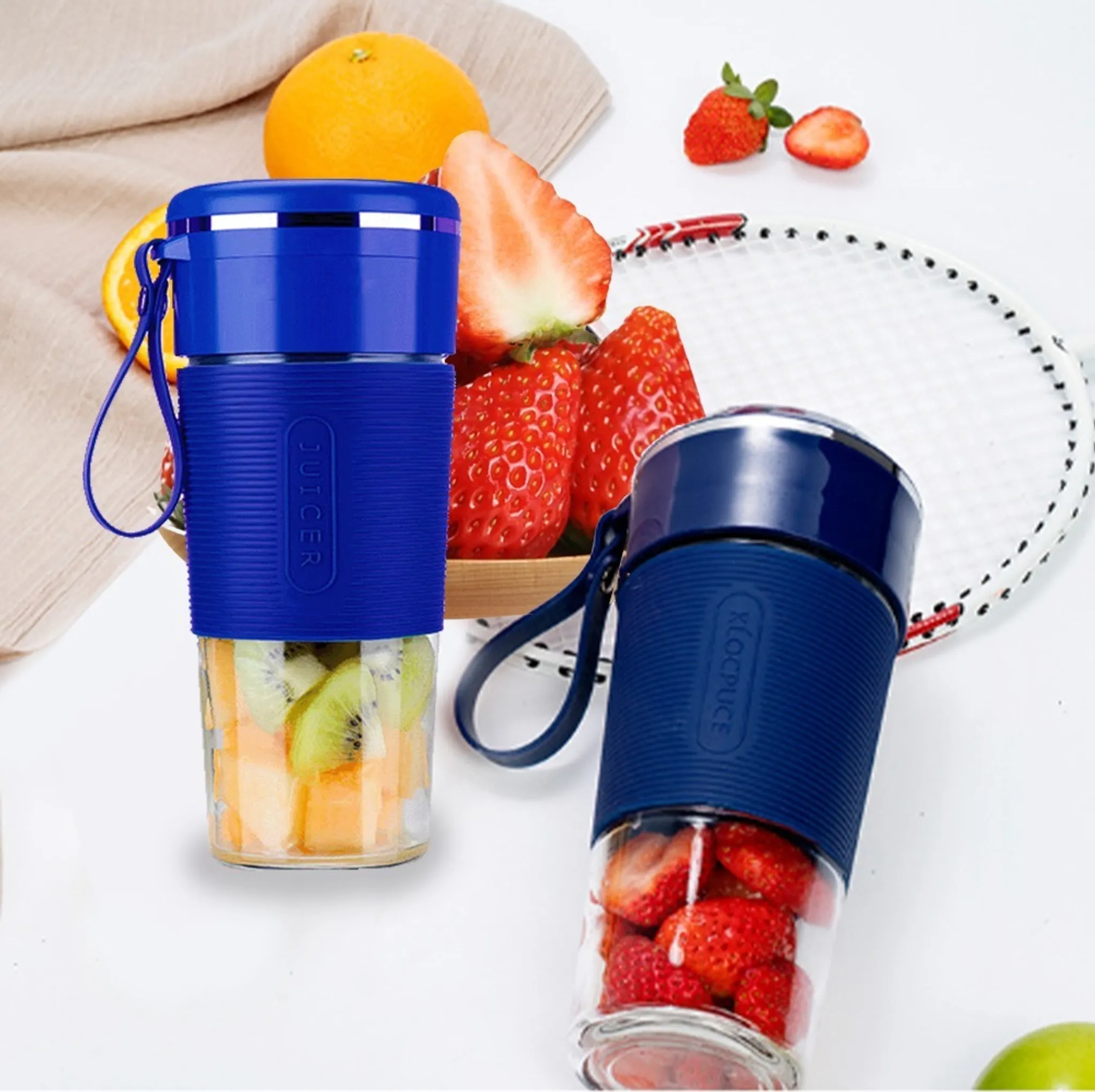 Zwei blaue Entsafter mit Früchten, Orangen, Erdbeeren und ein Tennisschläger liegen auf einer weißen Oberfläche.