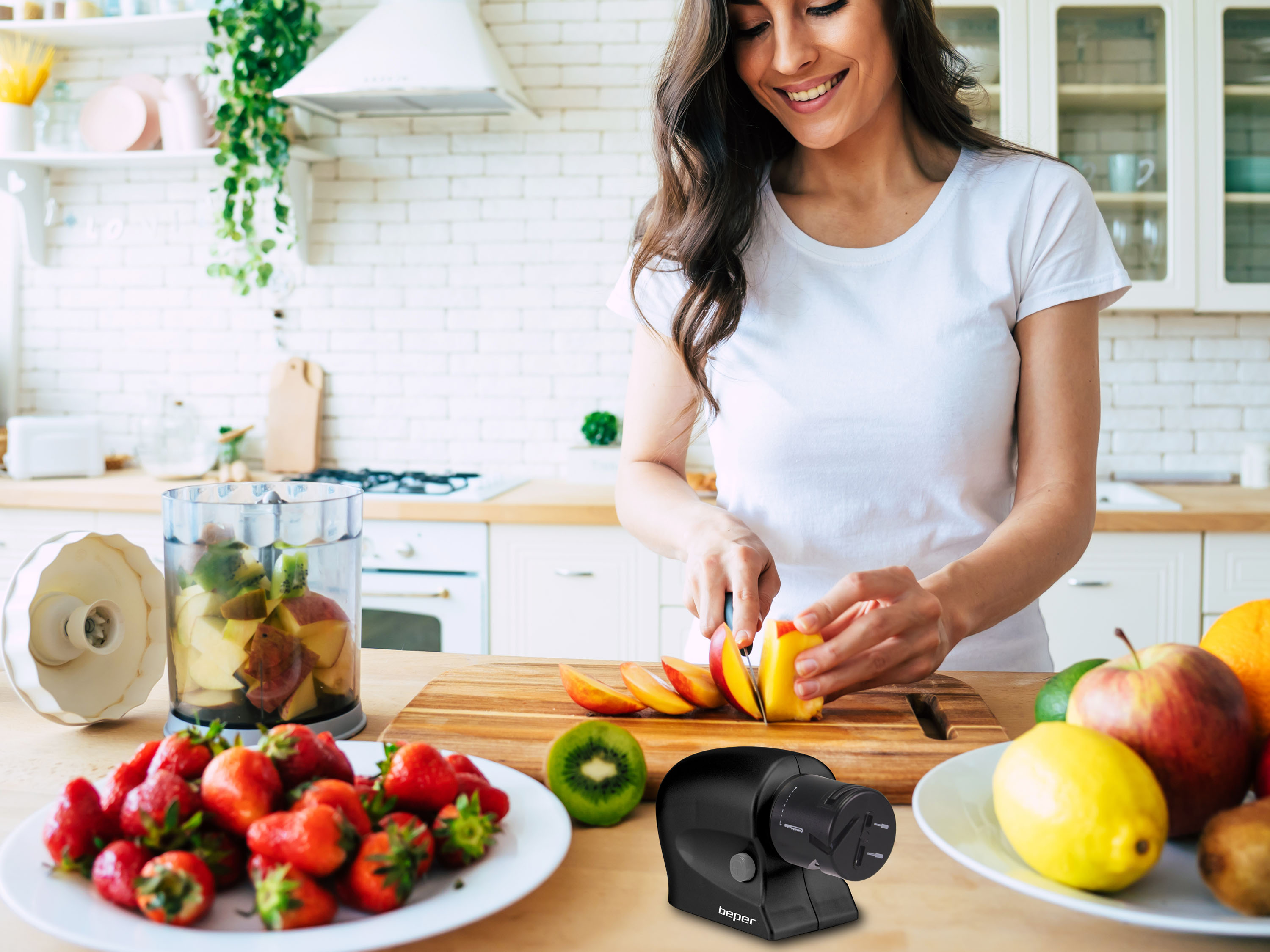 Donna taglia frutta in cucina. Fragole, kiwi e mele sul tavolo. Un frullatore e un affilacoltelli sono visibili.
