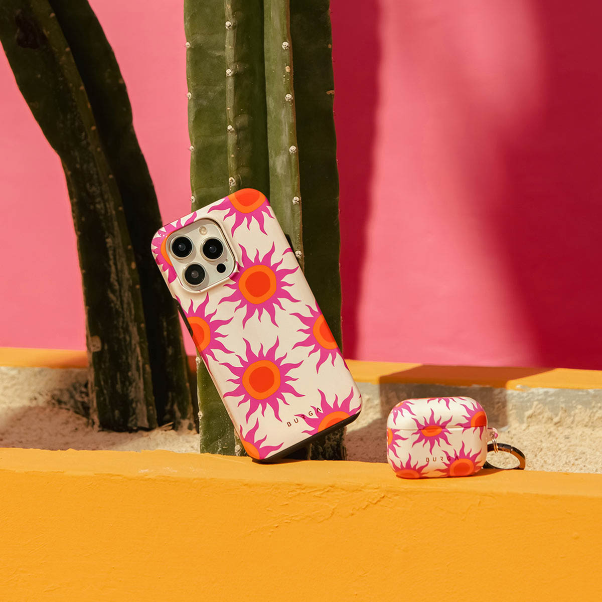 Een telefoonhoesje en een oortjesetui met een zonnebloempatroon. Ze staan naast een cactus met een roze achtergrond en een gele richel.