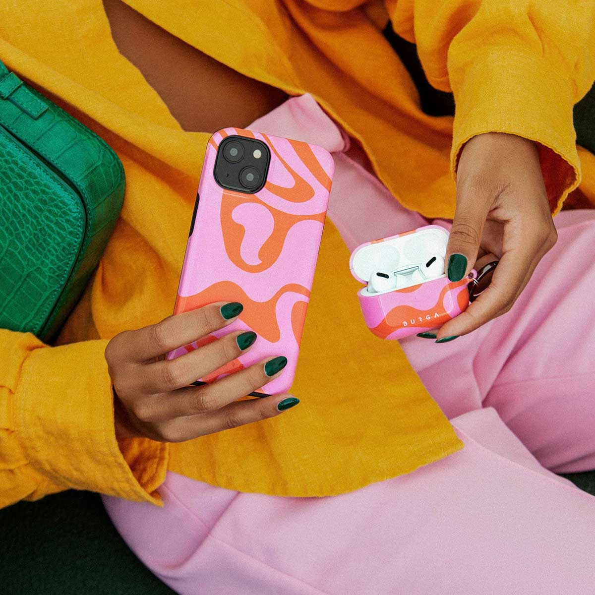 Vrouw houdt een telefoon en een oortelefoonhoesje vast met een roze en oranje wervelontwerp. Draagt een gele blouse, roze broek en groene nagellak.