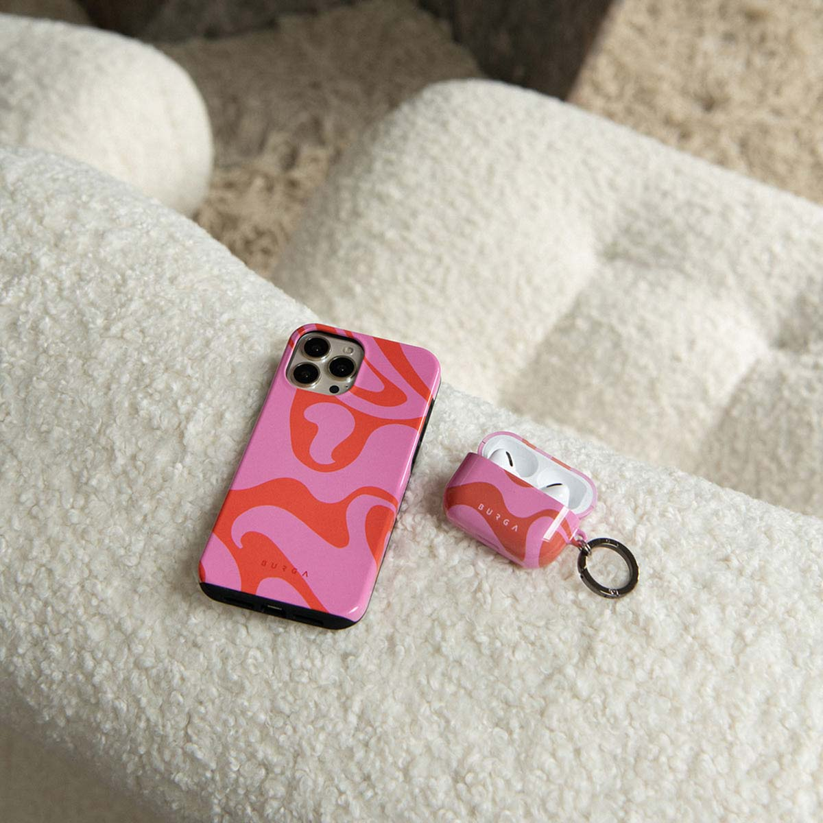 Roze telefoonhoesje en AirPod-hoesje met rode wervelingen op een wit, fuzzy oppervlak. Op de achtergrond een witte, fuzzy stoel.