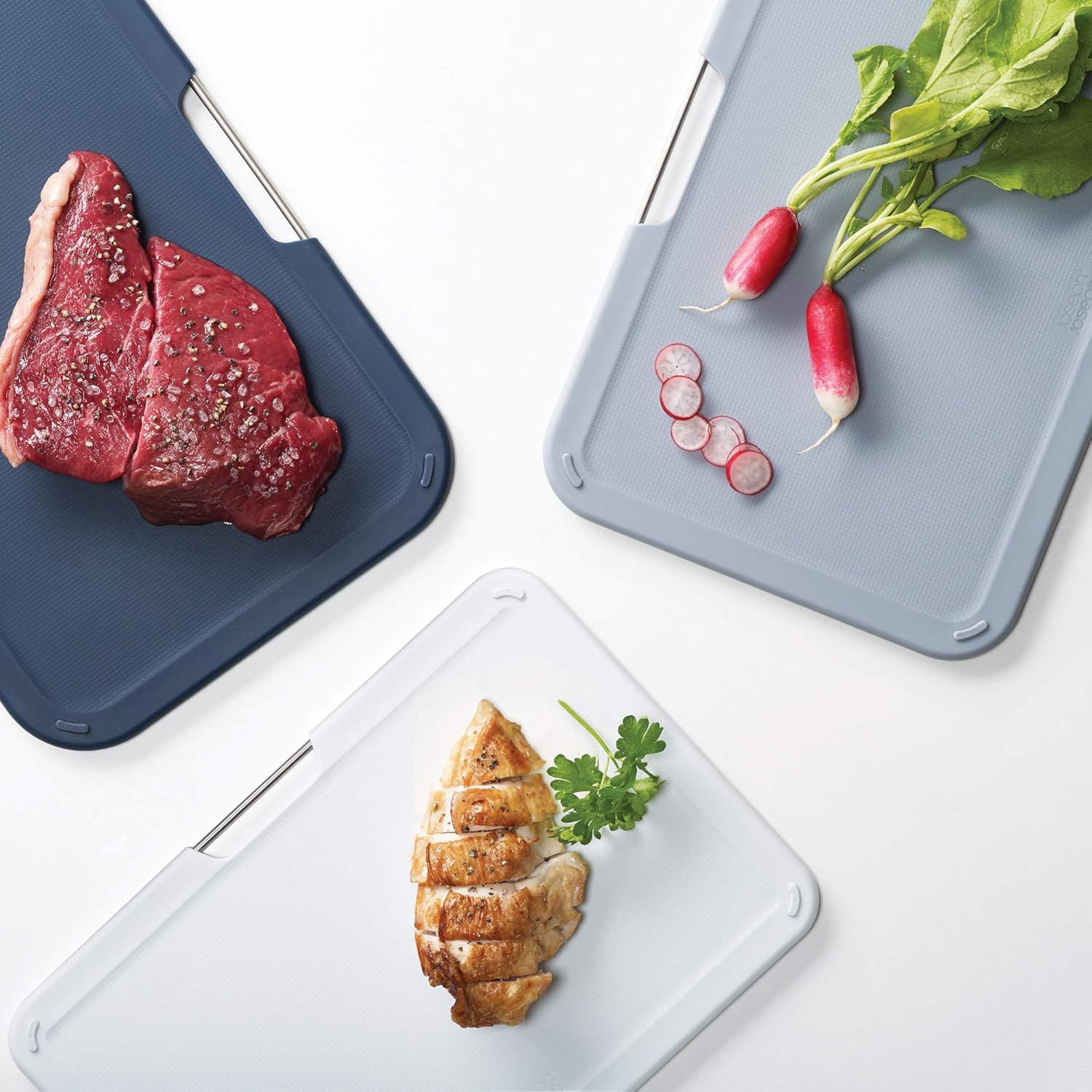 Drei Schneidebretter mit Essen: rohes Steak, Radieschenscheiben und gekochtes Hähnchen. Die Bretter sind verschiedenfarbig.
