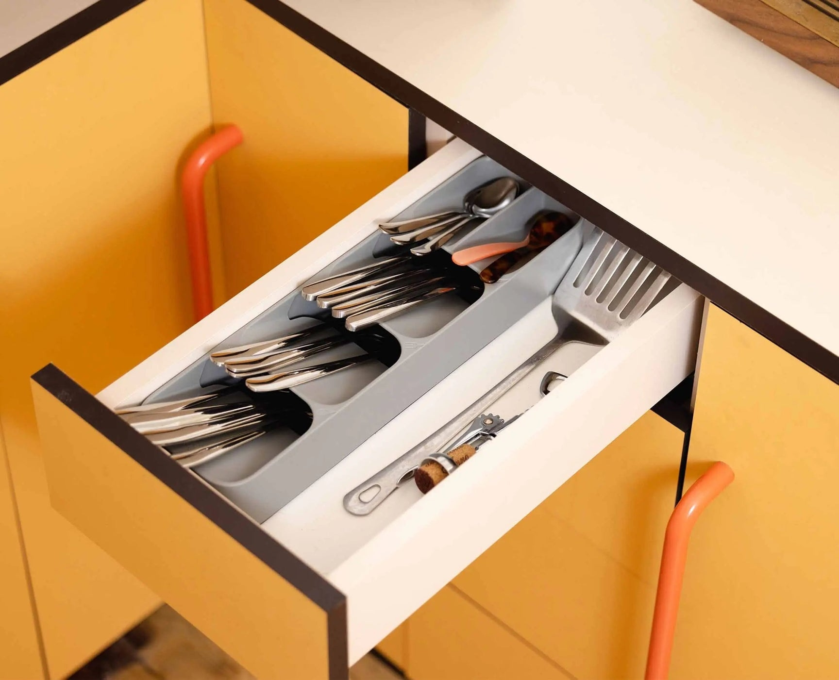 Cajón de cocina abierto con cubiertos. Tenedores, cucharas y un abridor de botellas visibles en el organizador gris. Tiradores naranjas y armarios amarillos.