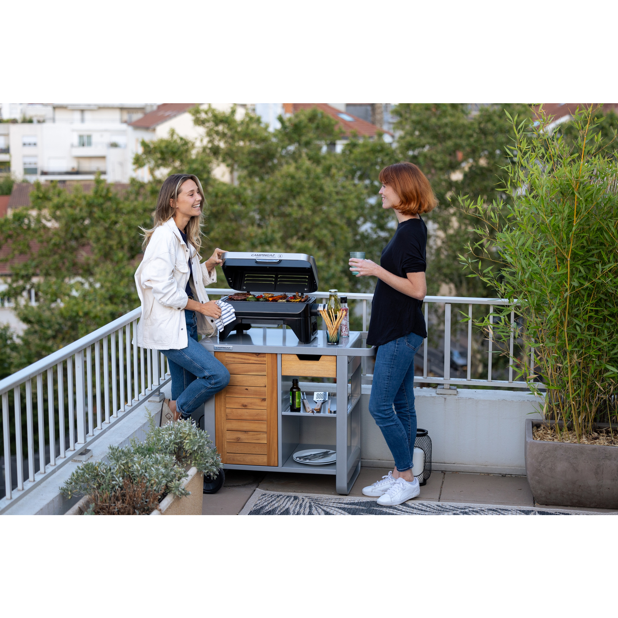 Zwei Frauen auf einem Balkon mit Grill. Eine öffnet den Grill, die andere hält ein Glas. Gebäude und Bäume im Hintergrund.