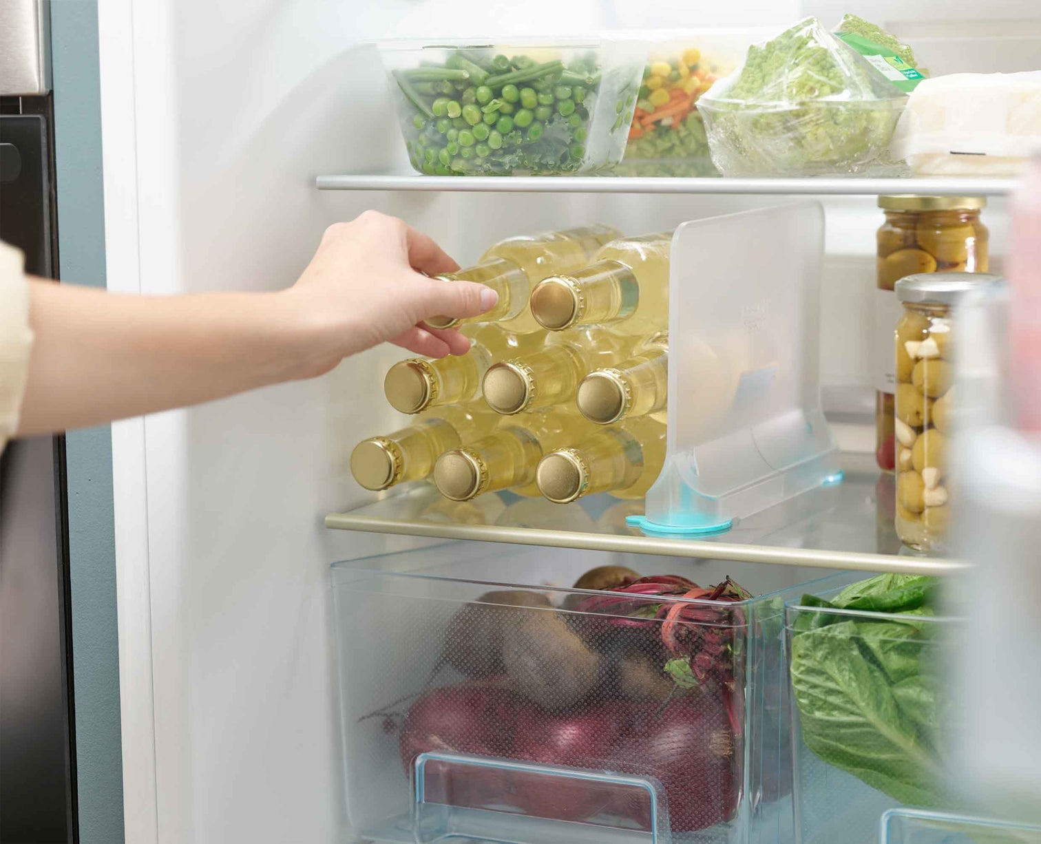 Ręka sięga do lodówki, wyjmując butelkę z stojaka na wino. Warzywa i słoiki są widoczne na półkach.