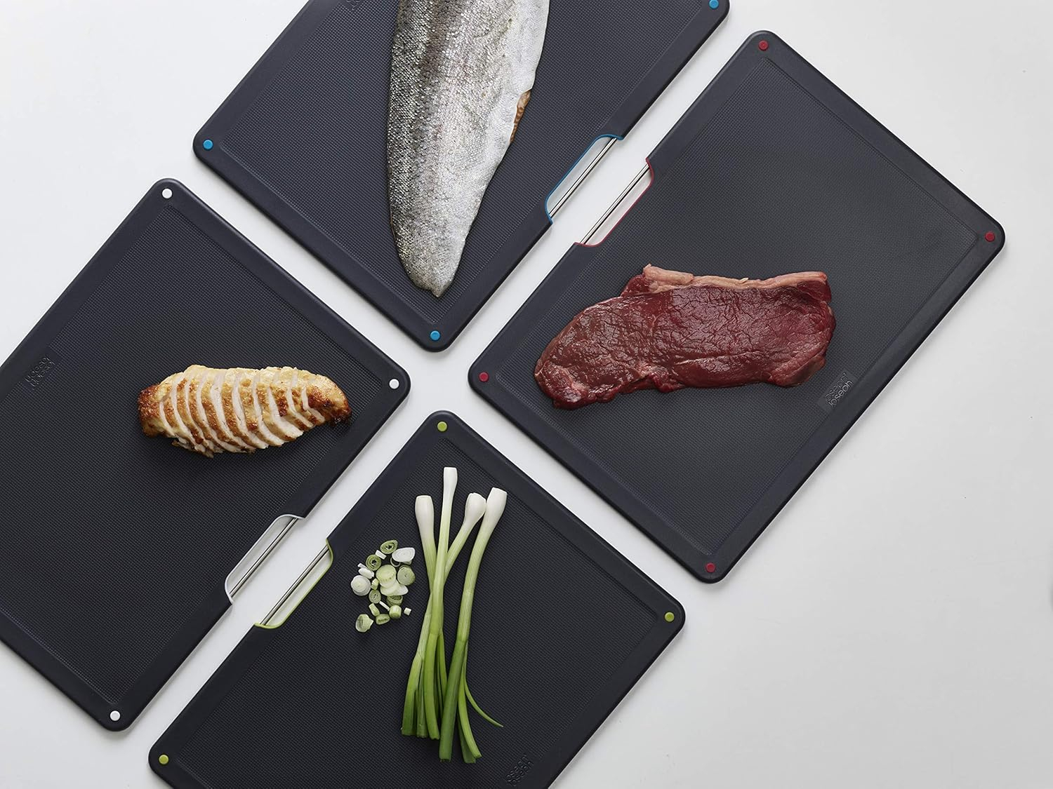 Cuatro tablas de cortar gris oscuro con alimentos: pescado, bistec, pollo y cebolletas. Las tablas tienen puntos de colores en las esquinas.