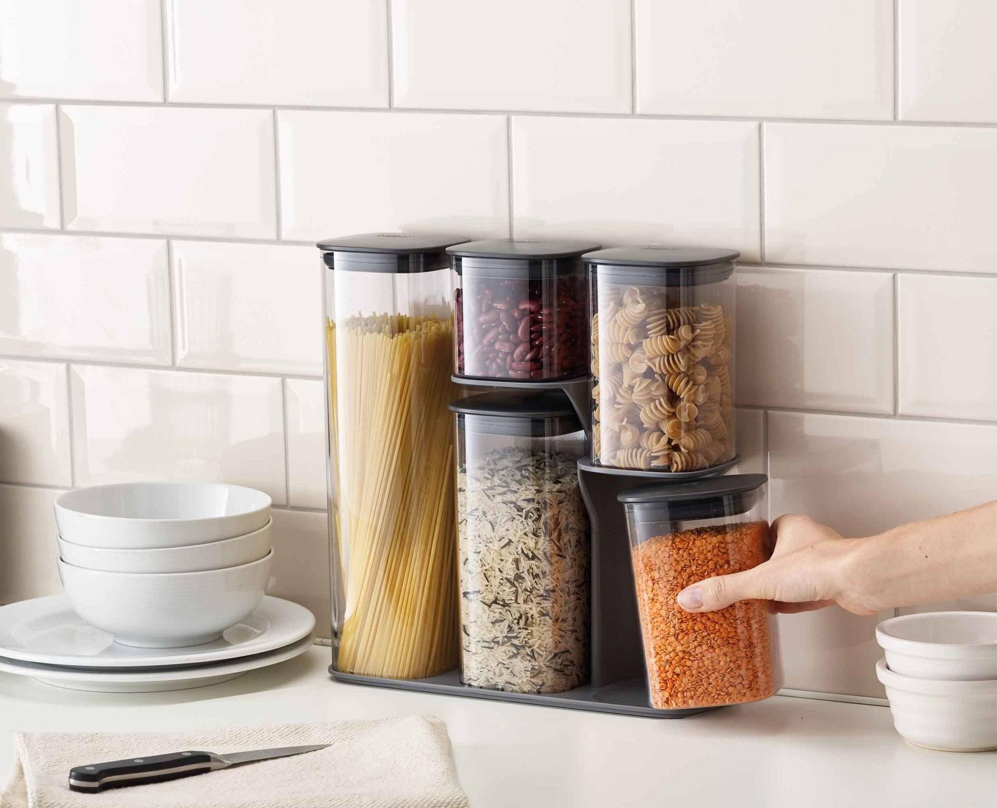Una escena de cocina con recipientes de almacenamiento llenos de pasta, frijoles y lentejas. Una mano alcanza un recipiente.