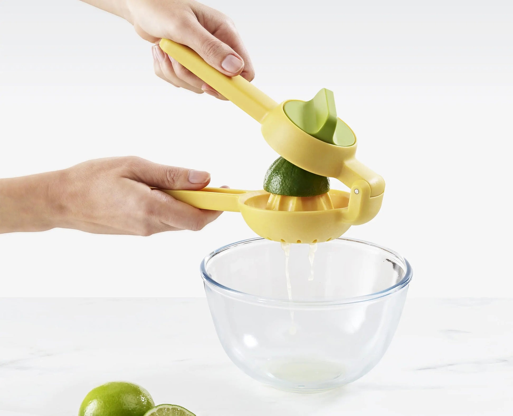 Una persona sprema un lime con uno spremiagrumi giallo sopra una ciotola trasparente. Il succo gocciola nella ciotola. Due lime sono sul tavolo.