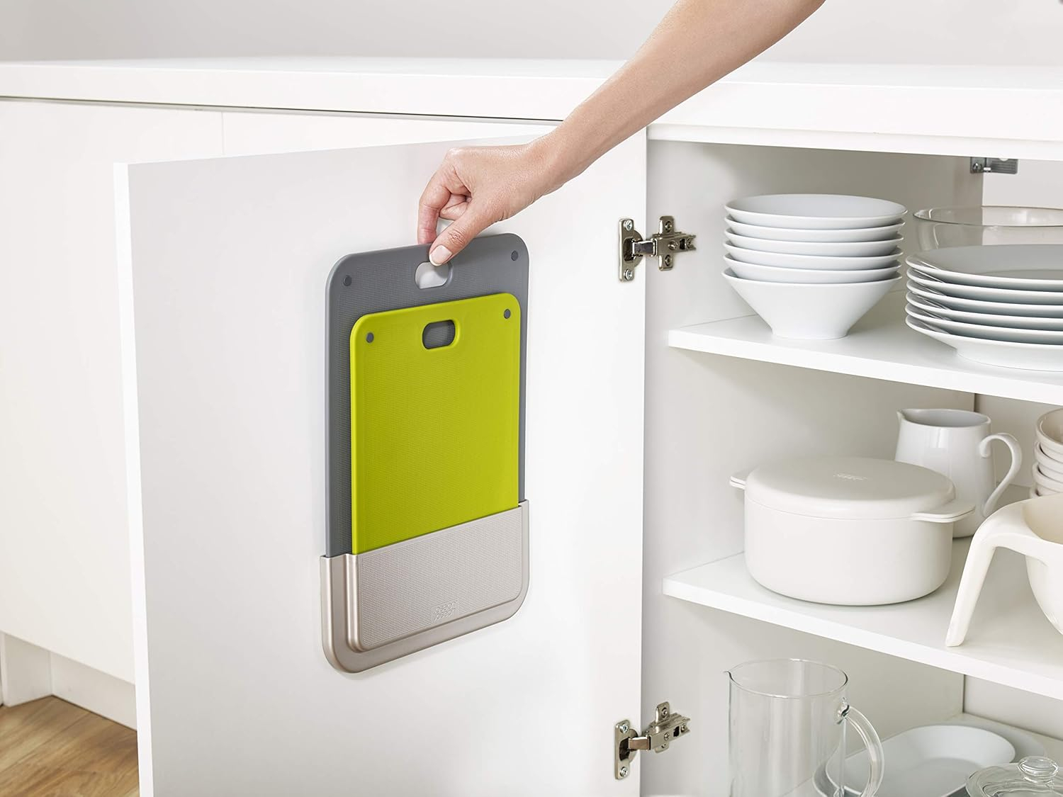 Una mano abre una puerta de armario blanco para revelar tablas de cortar apiladas. Una tabla gris y una verde están en un soporte de metal.