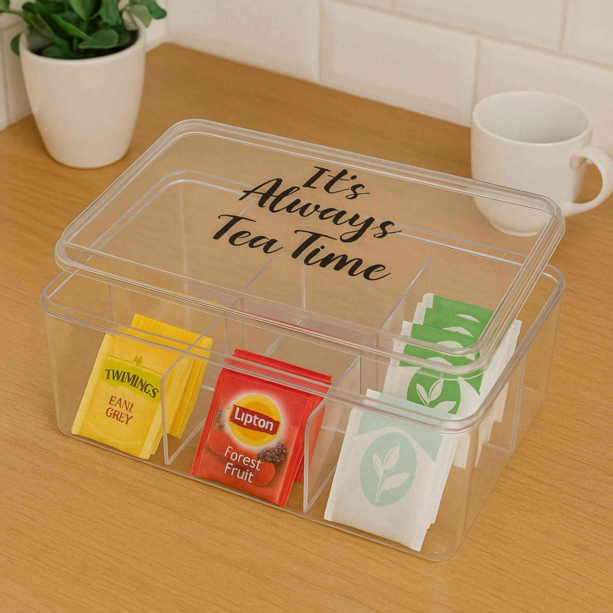 Una caja de té transparente con varias bolsitas de té, una taza y una pequeña planta sobre una superficie de madera.