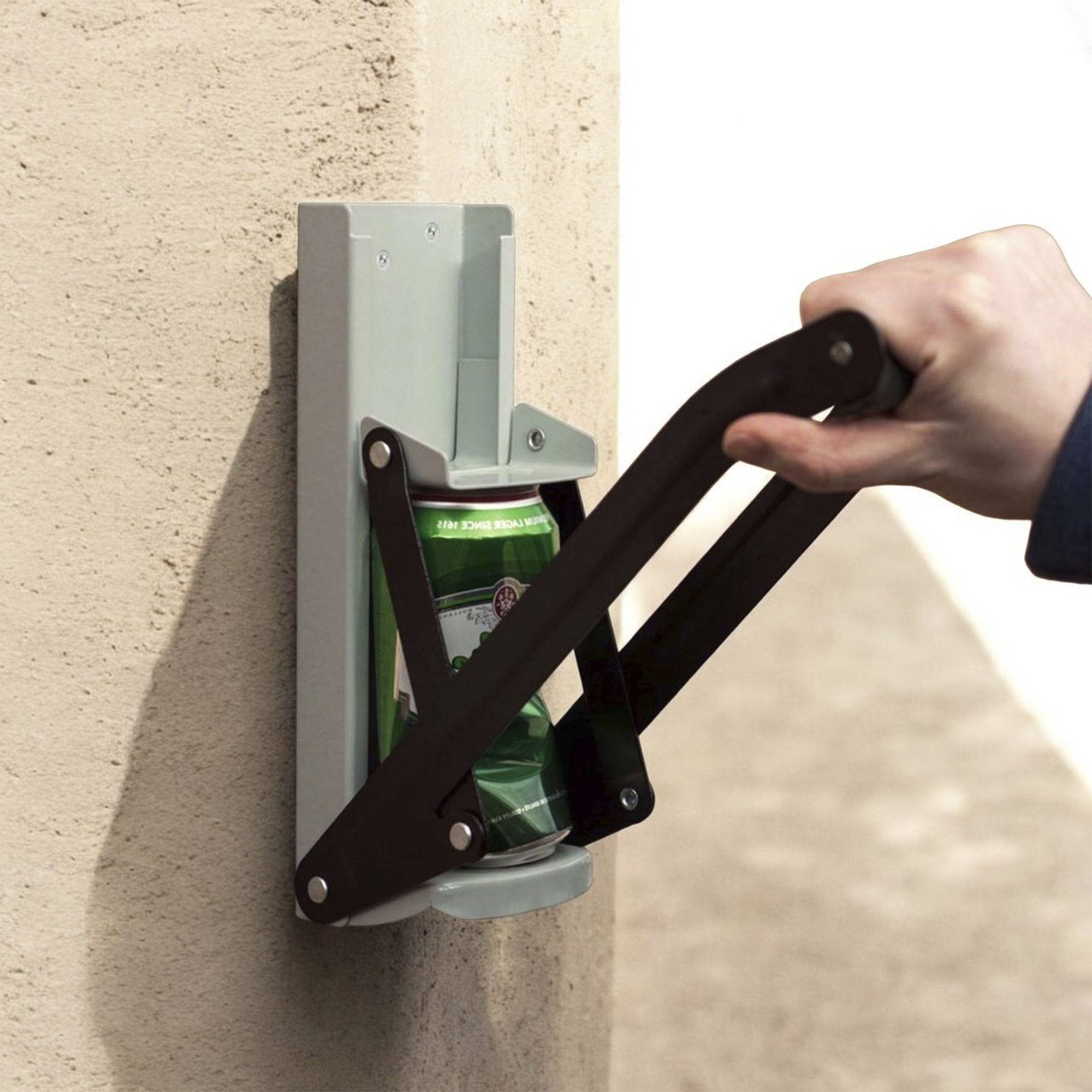 Una mano opera una trituradora de latas negra fijada a una pared. Una lata verde es aplastada. Pared beige.