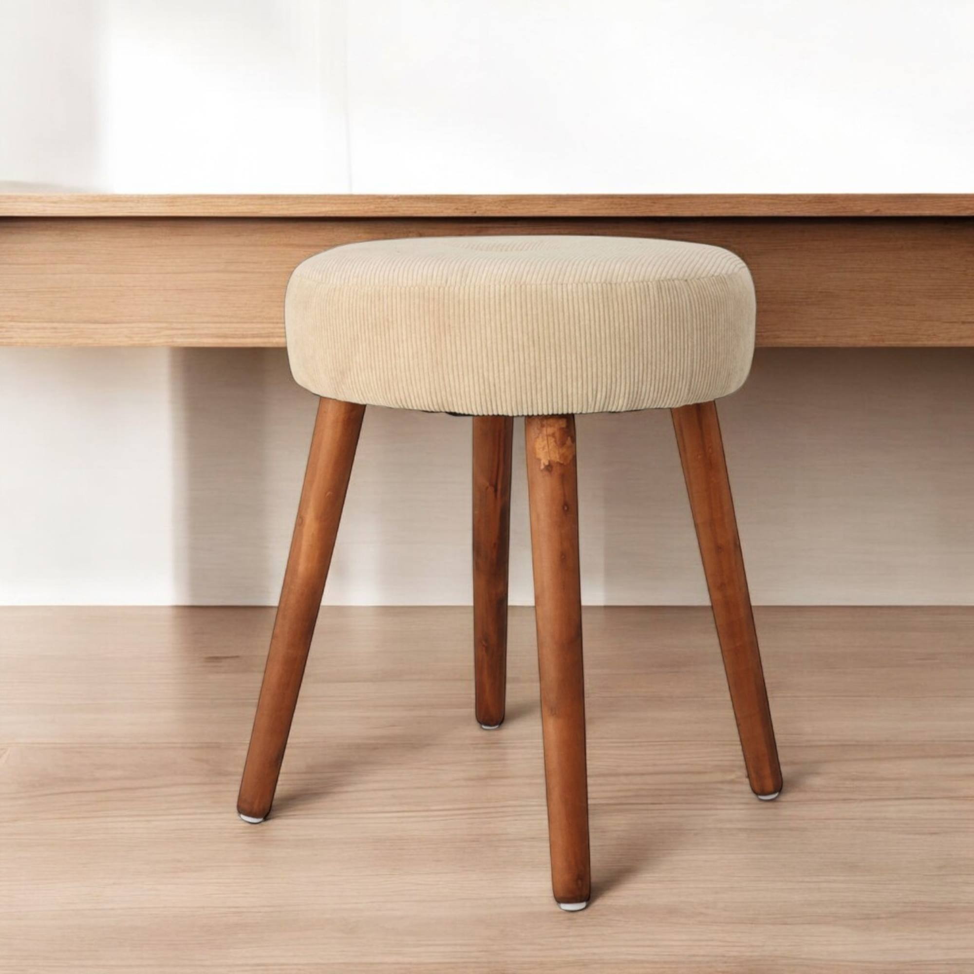 Un taburete de pana beige con patas de madera está sobre un piso de madera. Un escritorio de madera está en el fondo.