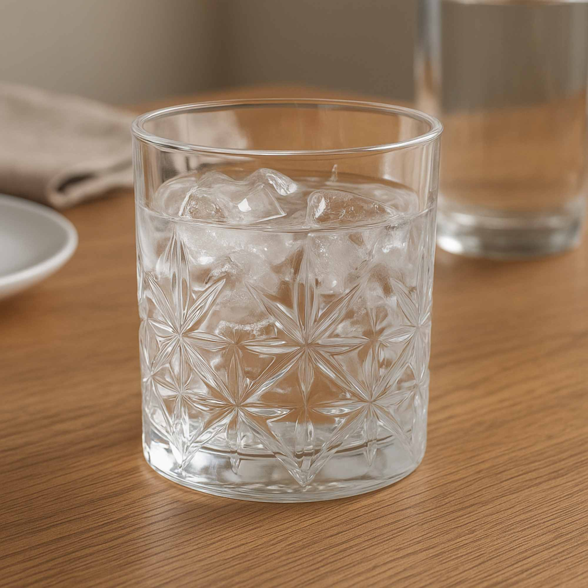 Un vaso estampado lleno de hielo y agua sobre una superficie de madera, con un vaso borroso al fondo.