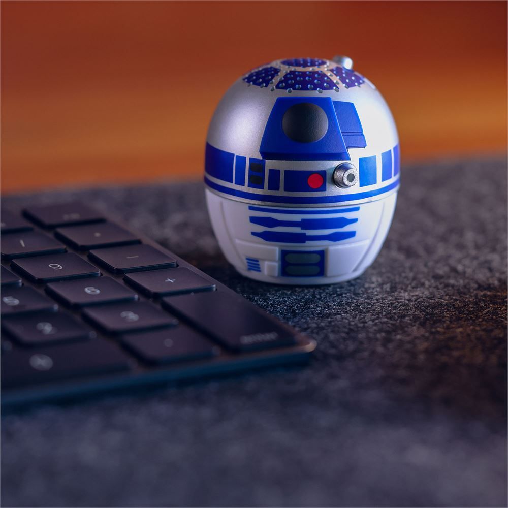 Bir R2-D2 oyuncağı, siyah bir klavyenin yanında, koyu gri bir yüzeyde duruyor. Arka plan bulanık turuncu ve kahverengi.