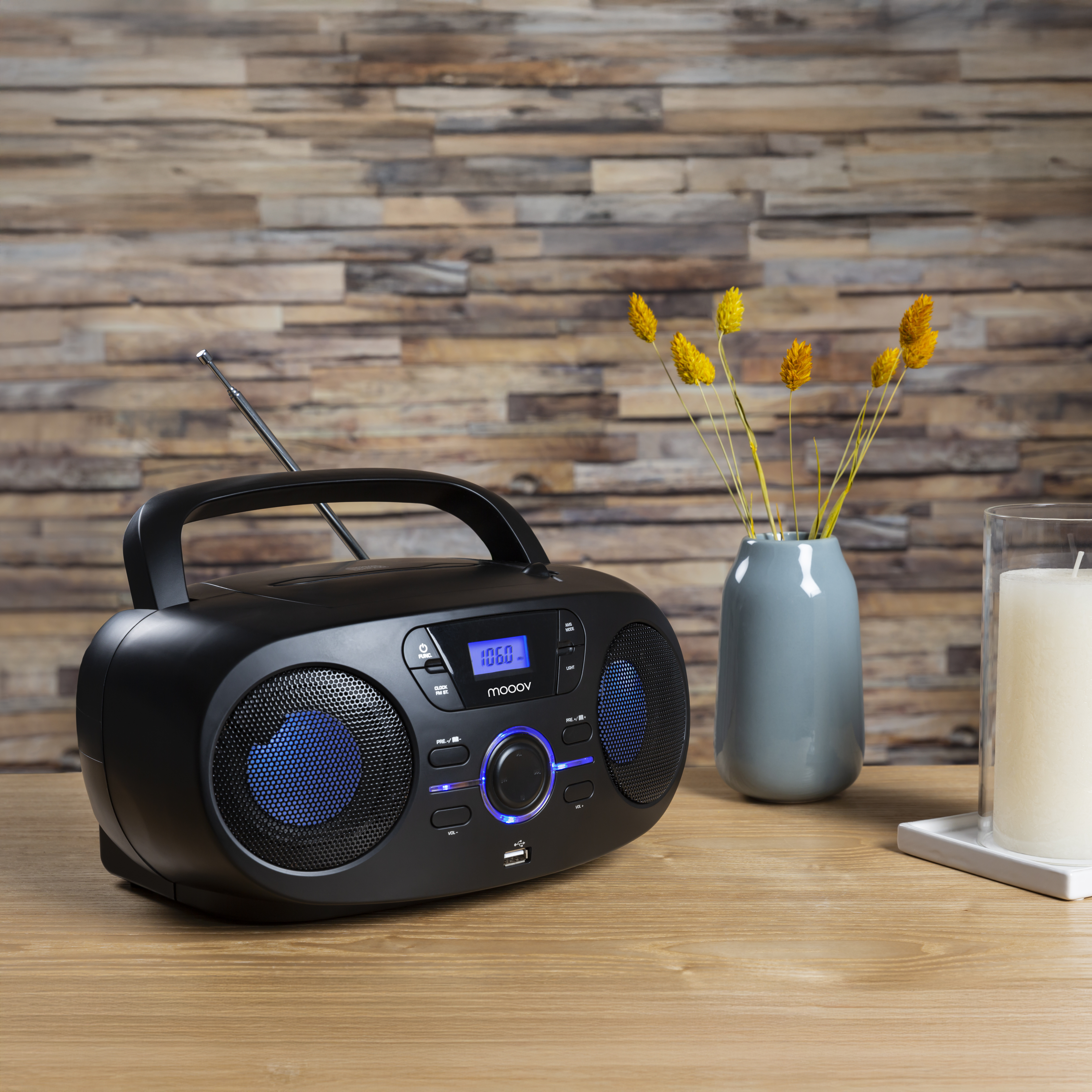 Una radio negra con altavoces azules está sobre una mesa de madera. Un jarrón con flores amarillas y una vela están cerca.