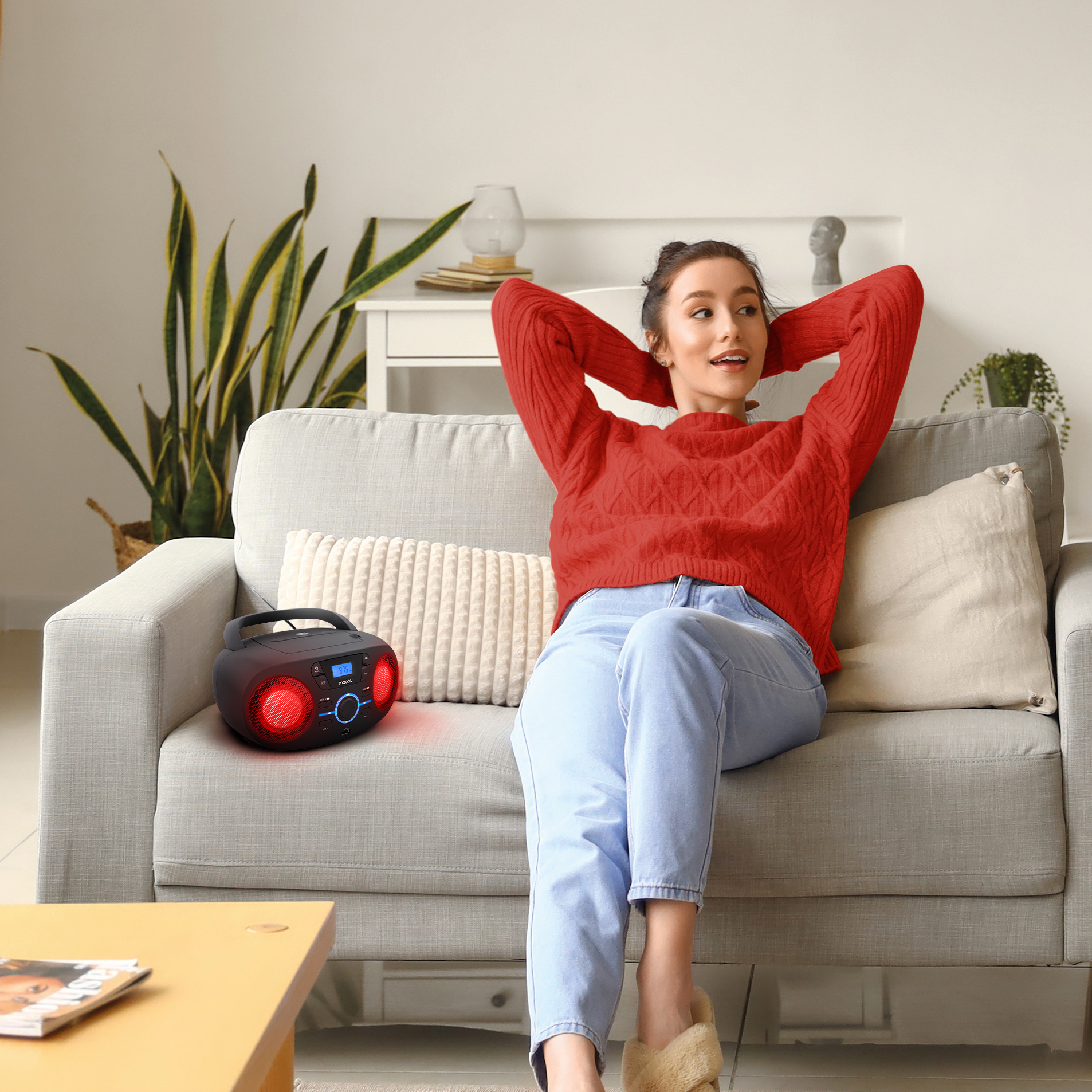 Mujer en suéter rojo en sofá, brazos detrás de la cabeza. Radio negro con luces rojas. Interior con plantas.