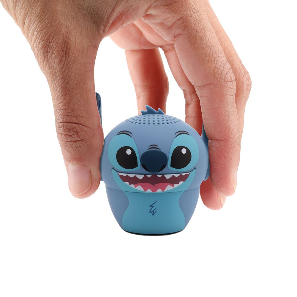Una mano tiene in mano un altoparlante a forma di Stitch. L'altoparlante blu ha occhi grandi, una bocca sorridente e piccole orecchie. Sfondo bianco.