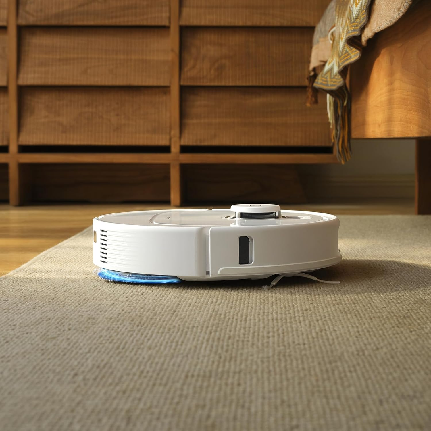 Een witte robotstofzuiger op een beige tapijt. Op de achtergrond een houten dressoir en een deel van een bed.
