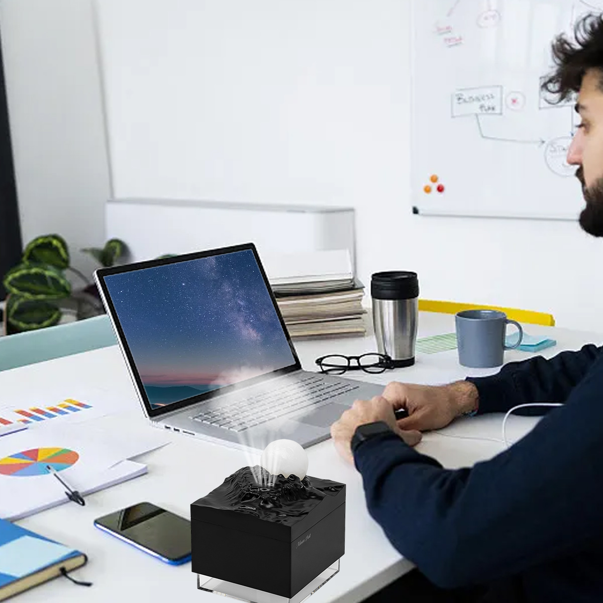 Ein Mann sitzt an einem Schreibtisch mit einem Laptop, der einen Sternenhimmel zeigt. Vorne steht eine schwarze Box mit einer Kugel. Weitere Gegenstände sind auf dem Schreibtisch.