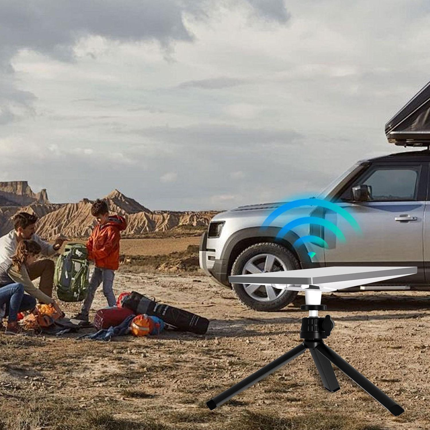 Famiglia in campeggio vicino a un SUV argentato. Un'antenna parabolica su un treppiede in primo piano. Montagne e cielo nuvoloso sullo sfondo.