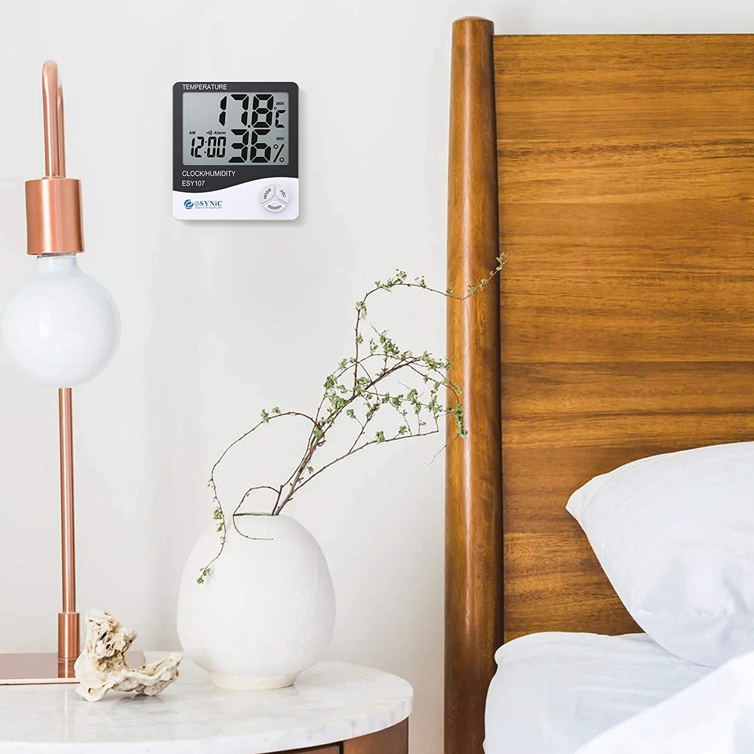 Ein Schlafzimmer mit einer Digitaluhr und einem Feuchtigkeitsmesser an einer weißen Wand. Eine weiße Vase mit grünen Stielen steht auf einem Tisch.