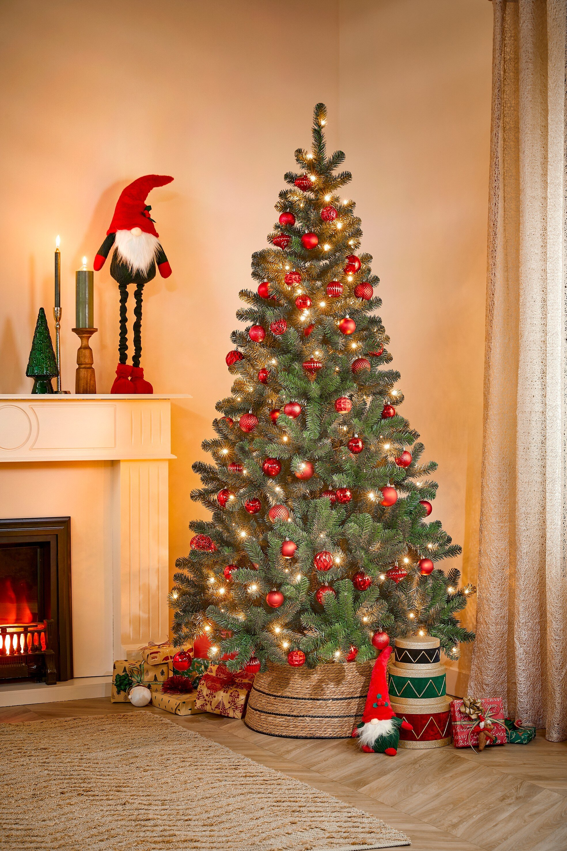 Un sapin de Noël décoré avec des ornements et des lumières rouges. Des cadeaux et des décorations entourent l'arbre, près d'une cheminée.
