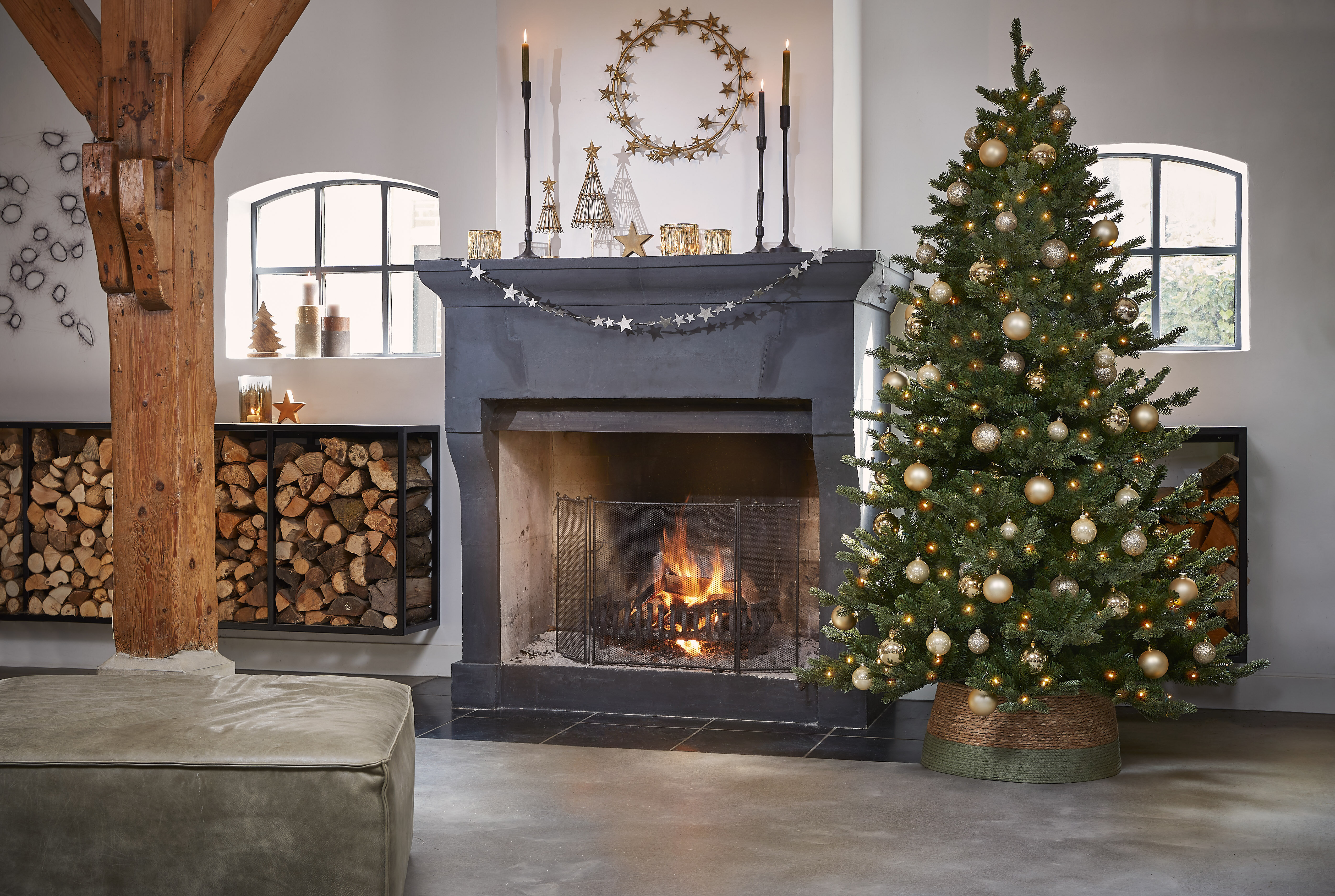 Un sapin de Noël décoré dans une pièce avec une cheminée et un rangement à bois. Des ornements et des lumières.
