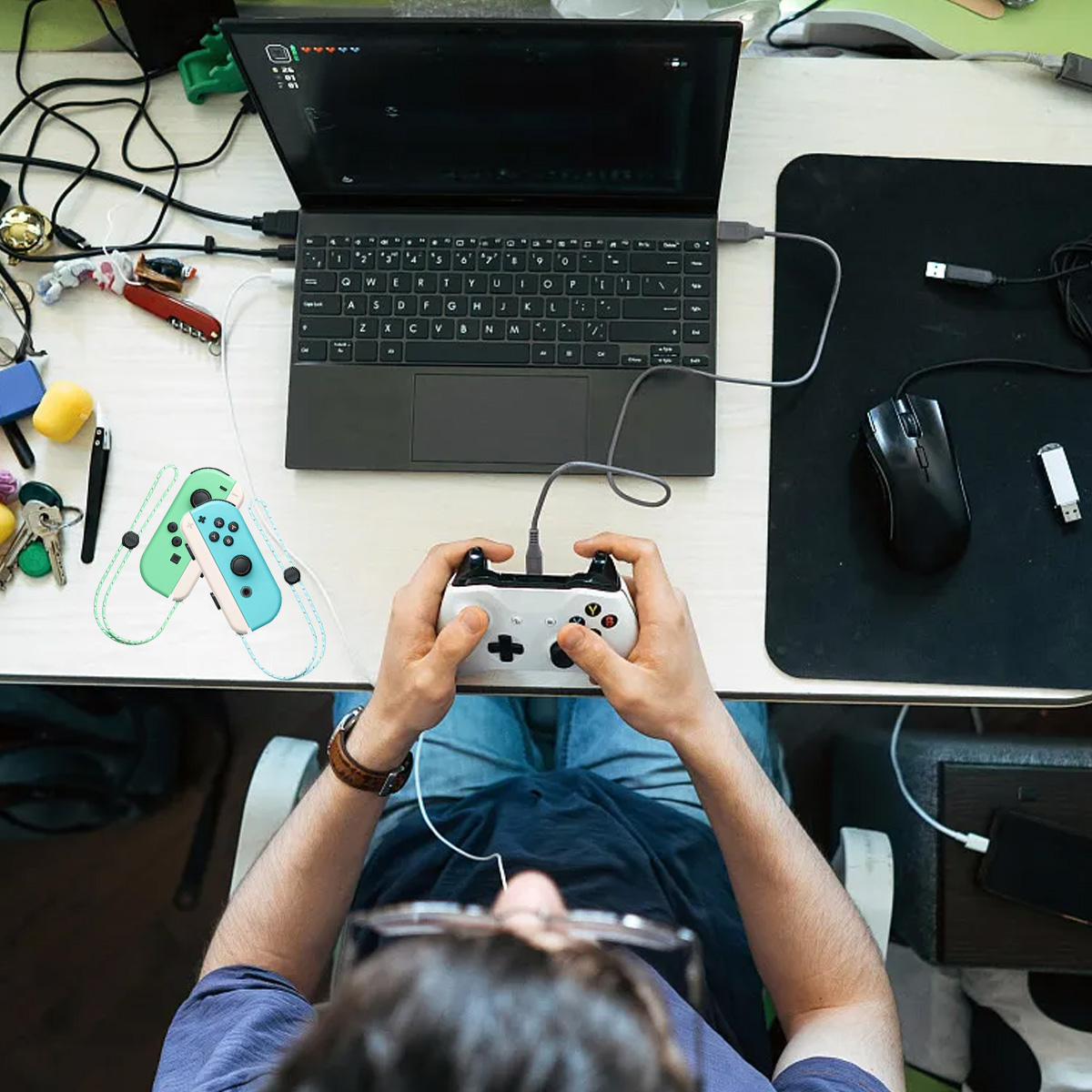 Eine Person hält einen weißen Gamecontroller, der mit einem Laptop verbunden ist. Maus und USB-Stick liegen auf einer schwarzen Matte. Switch-Controller liegen auf dem Schreibtisch.