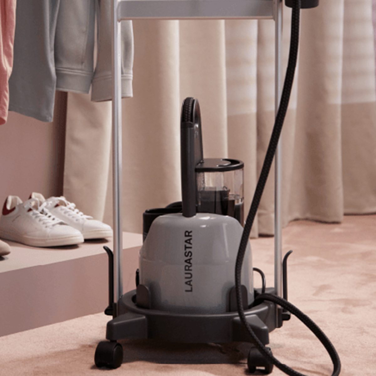 A gray Laurastar steamer, black cord, white sneakers, clothes on rack, and beige curtain in the background, set on a pink carpet.