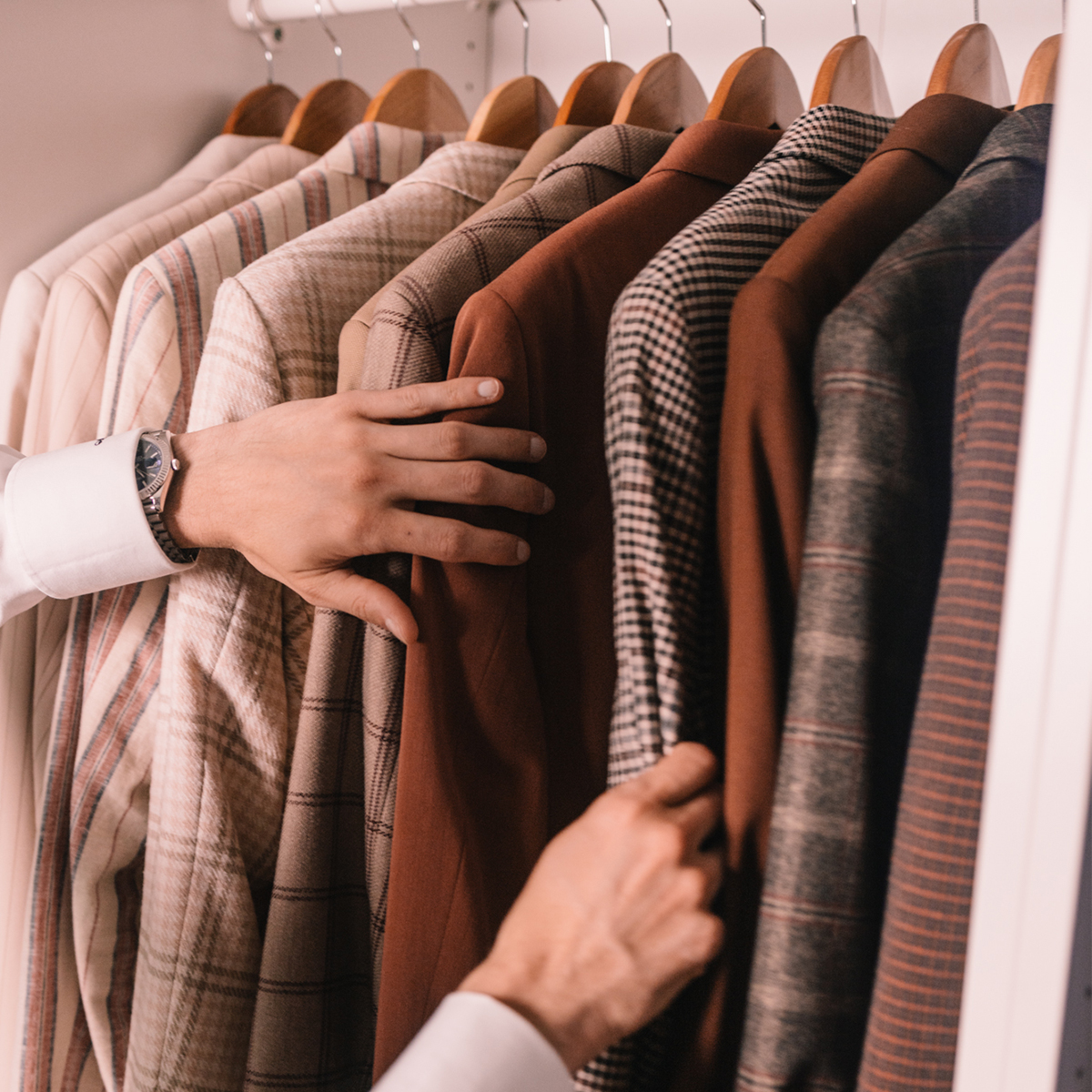 Hands selecting a jacket from a closet filled with various patterned suits. A watch is visible on the wrist.