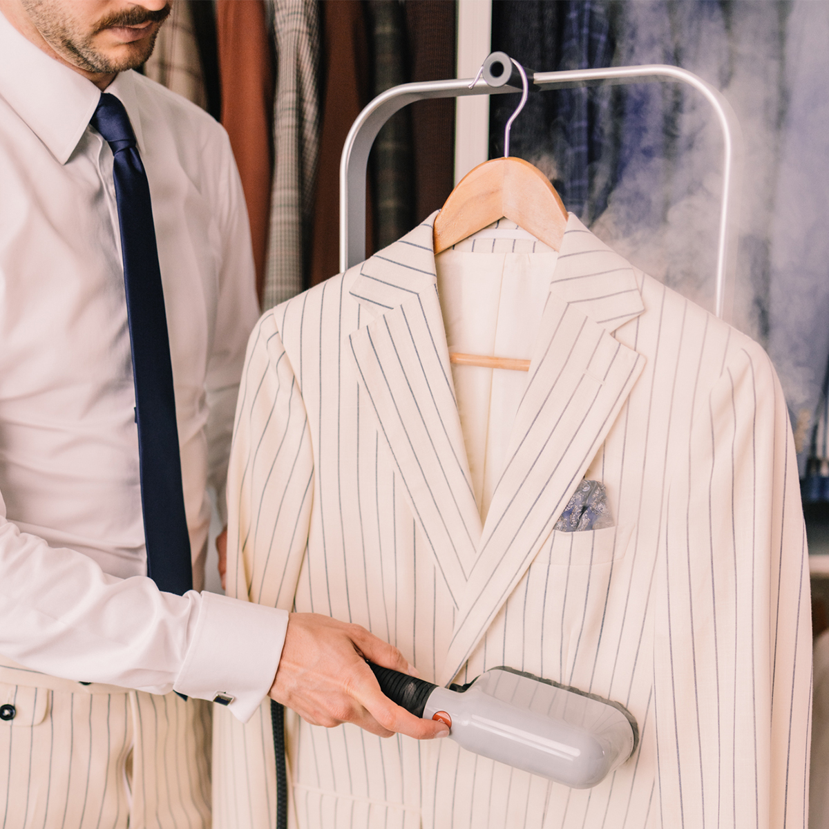 A man is steaming a cream striped suit jacket. The jacket hangs on a wooden hanger with steam rising. The man wears a white shirt and tie.