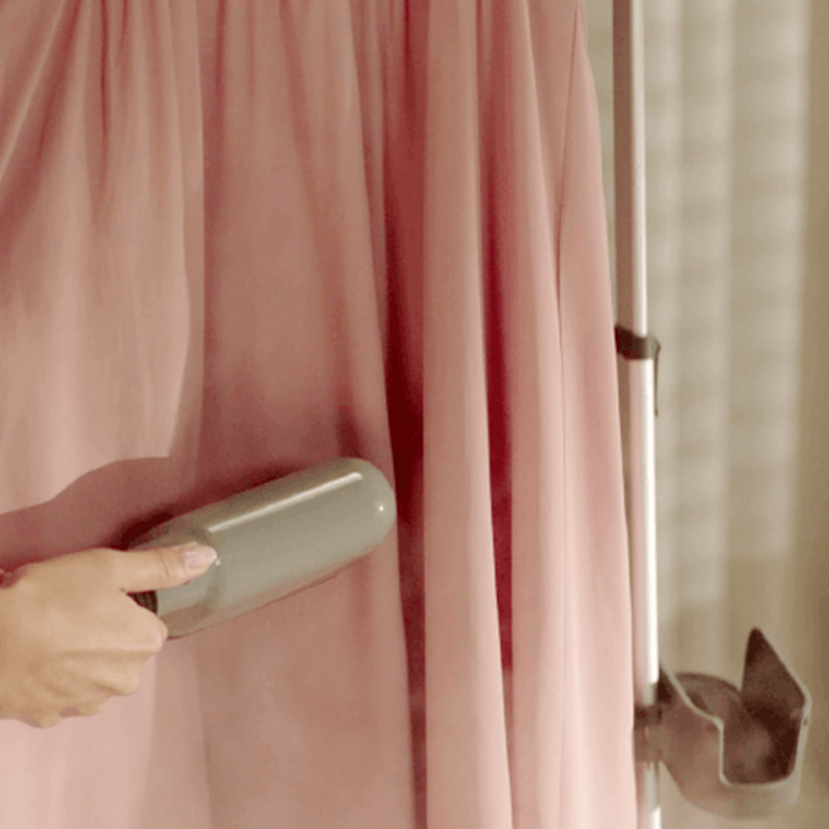 A hand using a gray steamer on a pink curtain. A metal bar and a holder for the steamer are on the right.